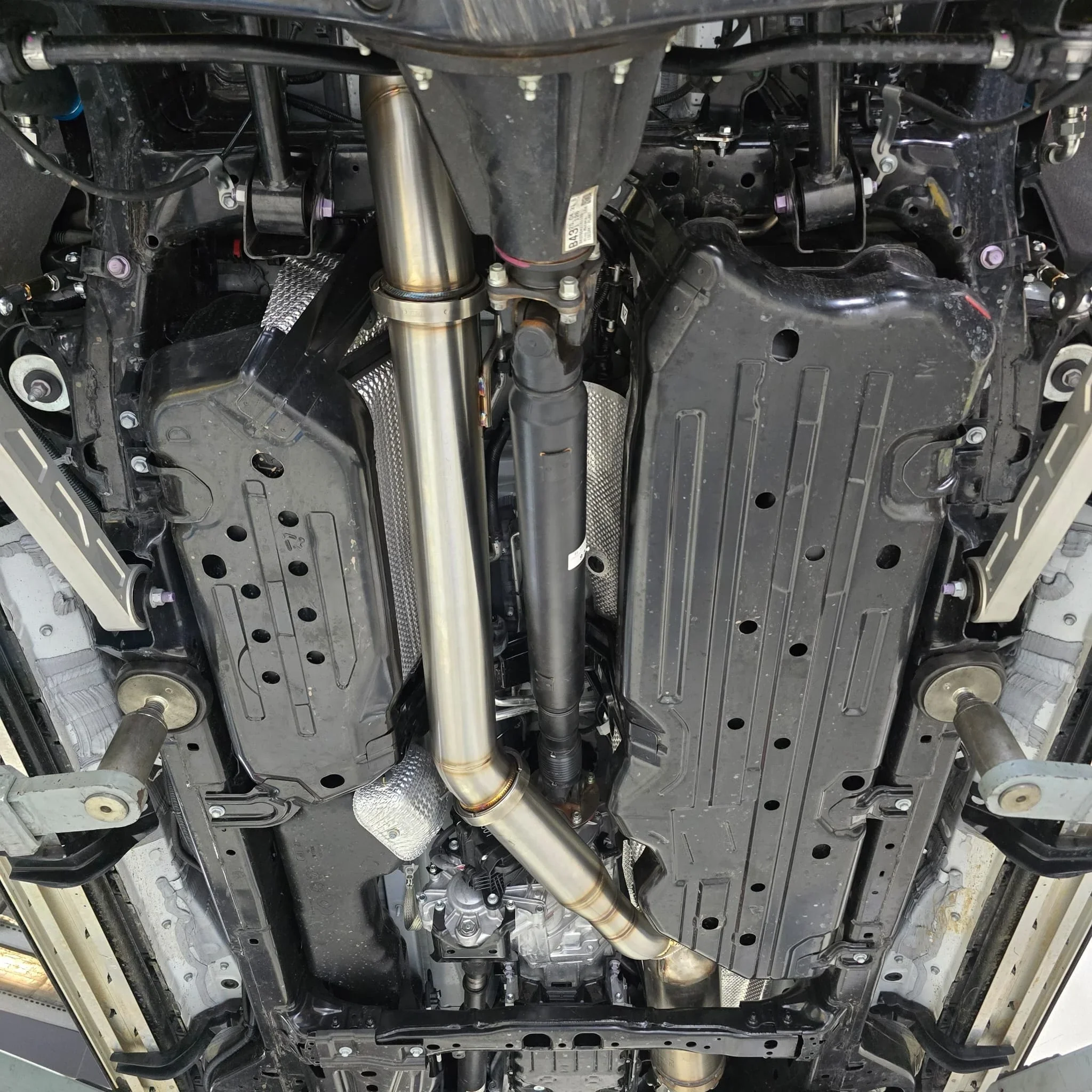 Underside of a car showing the engine, transmission, exhaust pipes, and various mechanical components.