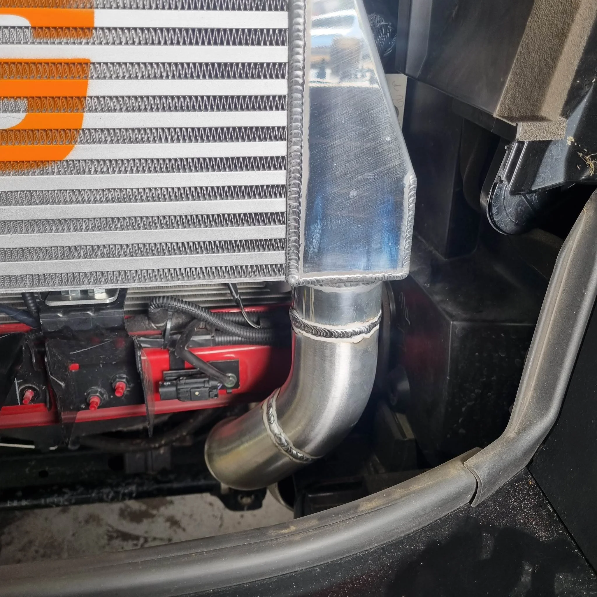 Close-up of a vehicle's engine showing a metal intercooler, polished metal pipe, and parts of the engine block with red accents.