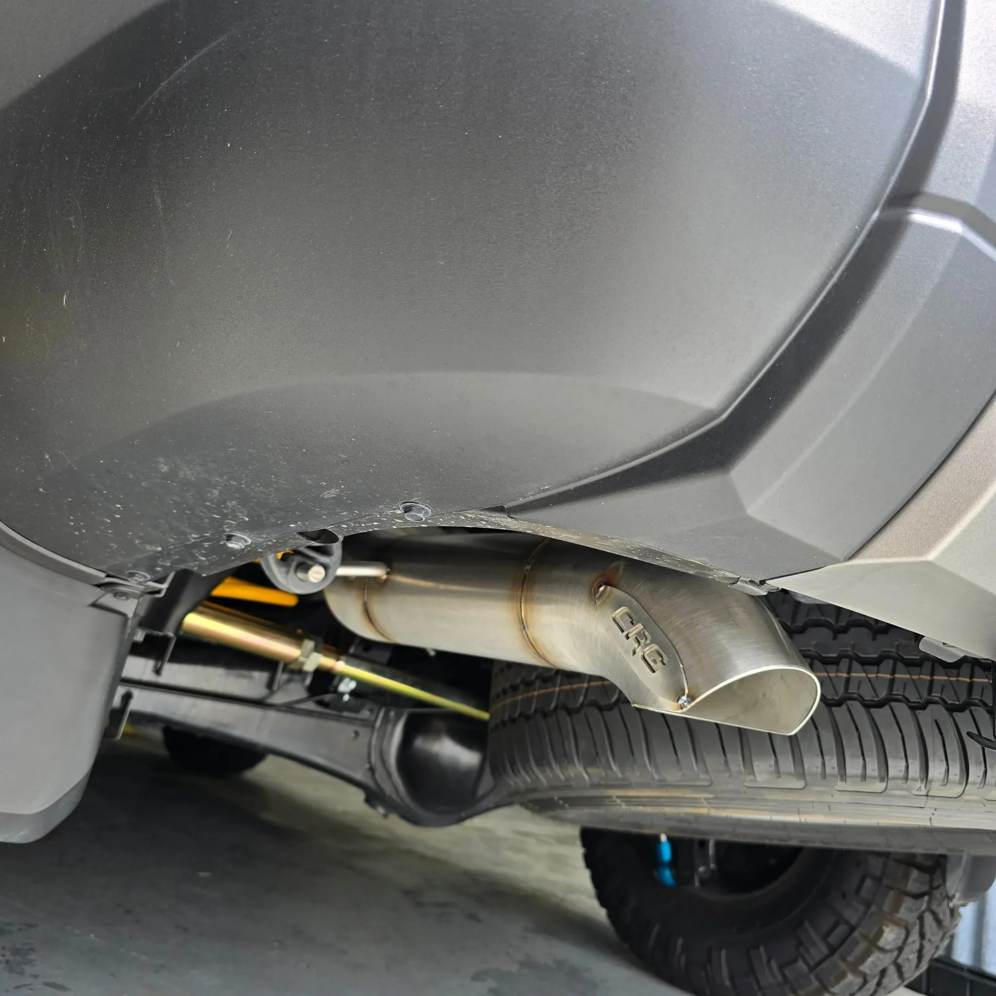 Close-up of a car's undercarriage showing a shiny exhaust pipe, part of the rear suspension, tire, and chassis.