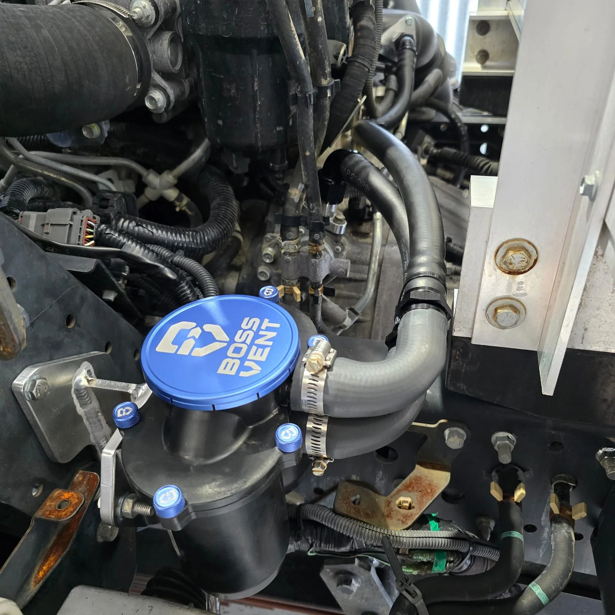 Close-up of a vehicle engine with a blue BOSS Vent cap on a component, numerous hoses, wires, and metal parts visible.