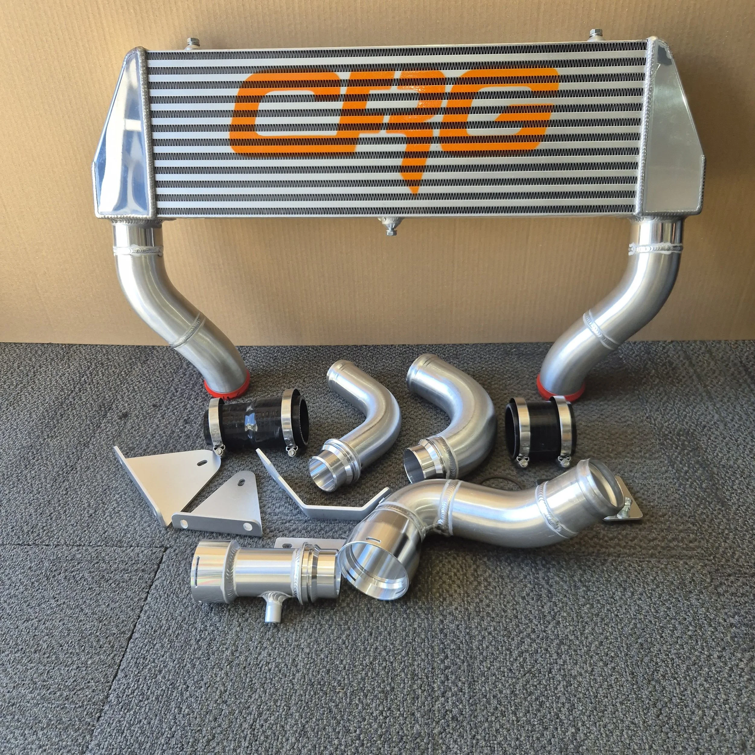 A car intercooler with orange logo and several shiny silver metal pipes and black couplings