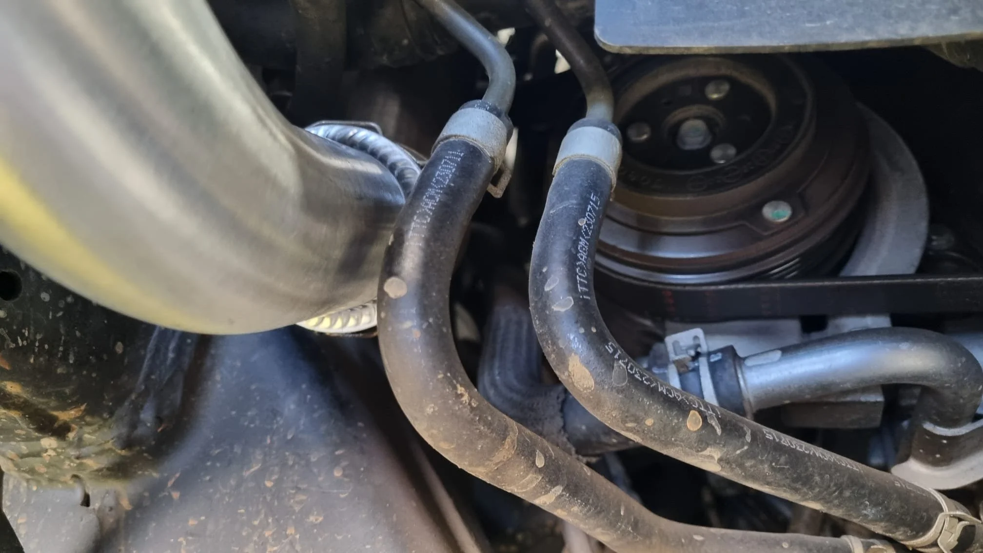 Close-up view of a car engine showing rubber hoses, metal pipes, a belt pulley, and part of the engine block.
