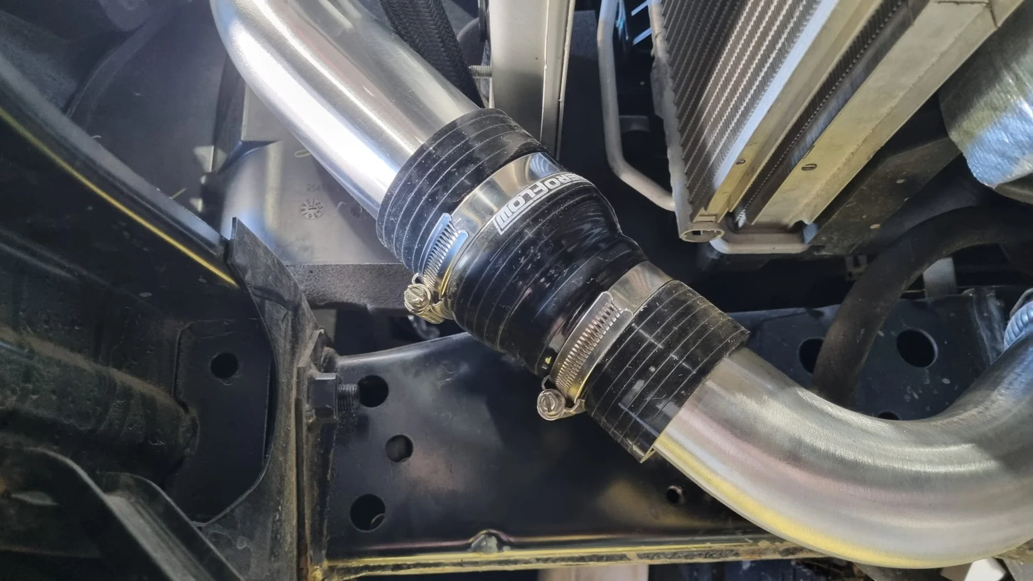 Close-up of a car's suspension system showing a shiny metal coilover with black rubber and metal clamps, and a portion of the radiator or cooling system in the background.