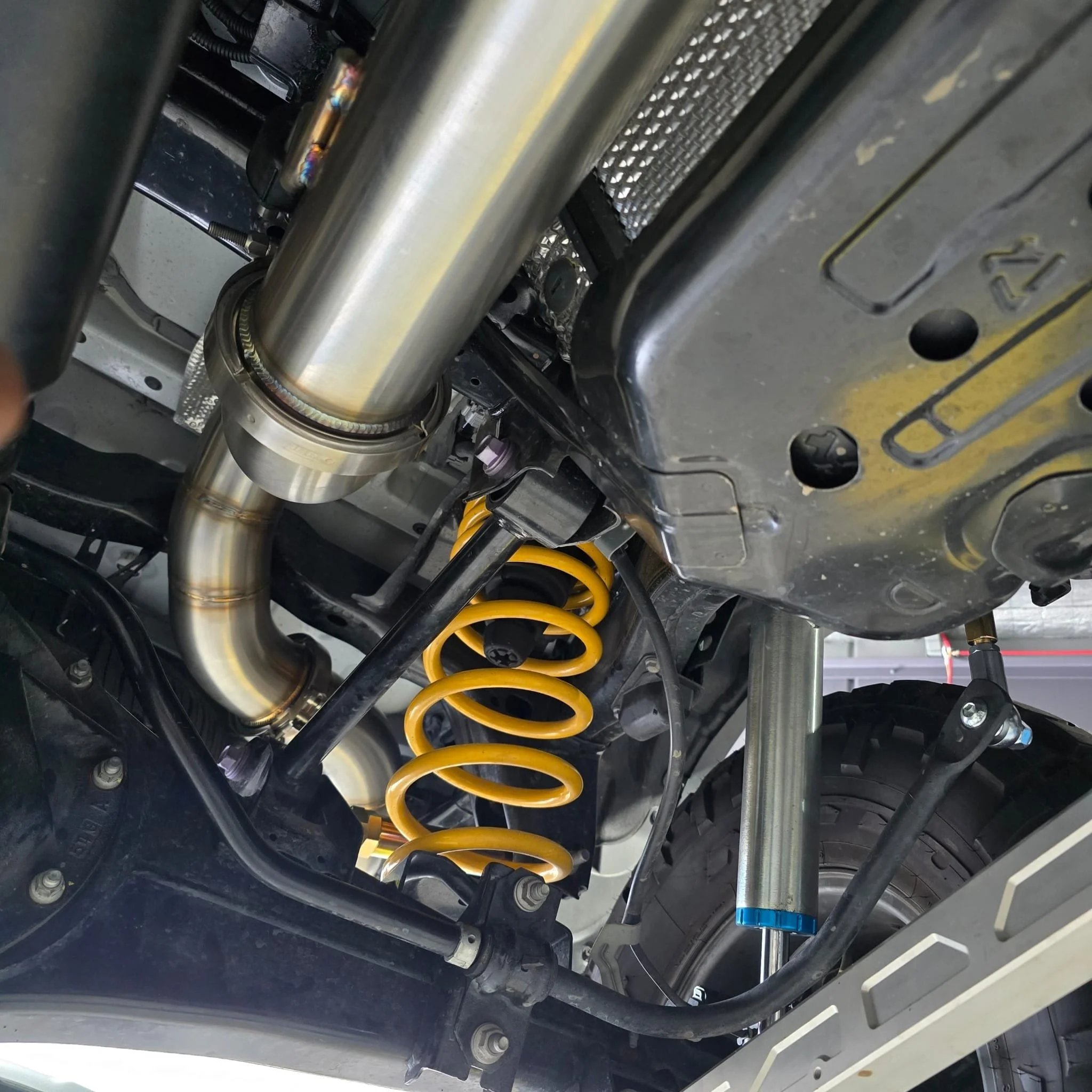 Close-up view of a vehicle's suspension system showing a yellow coil spring, shock absorber, and parts of the chassis and undercarriage.