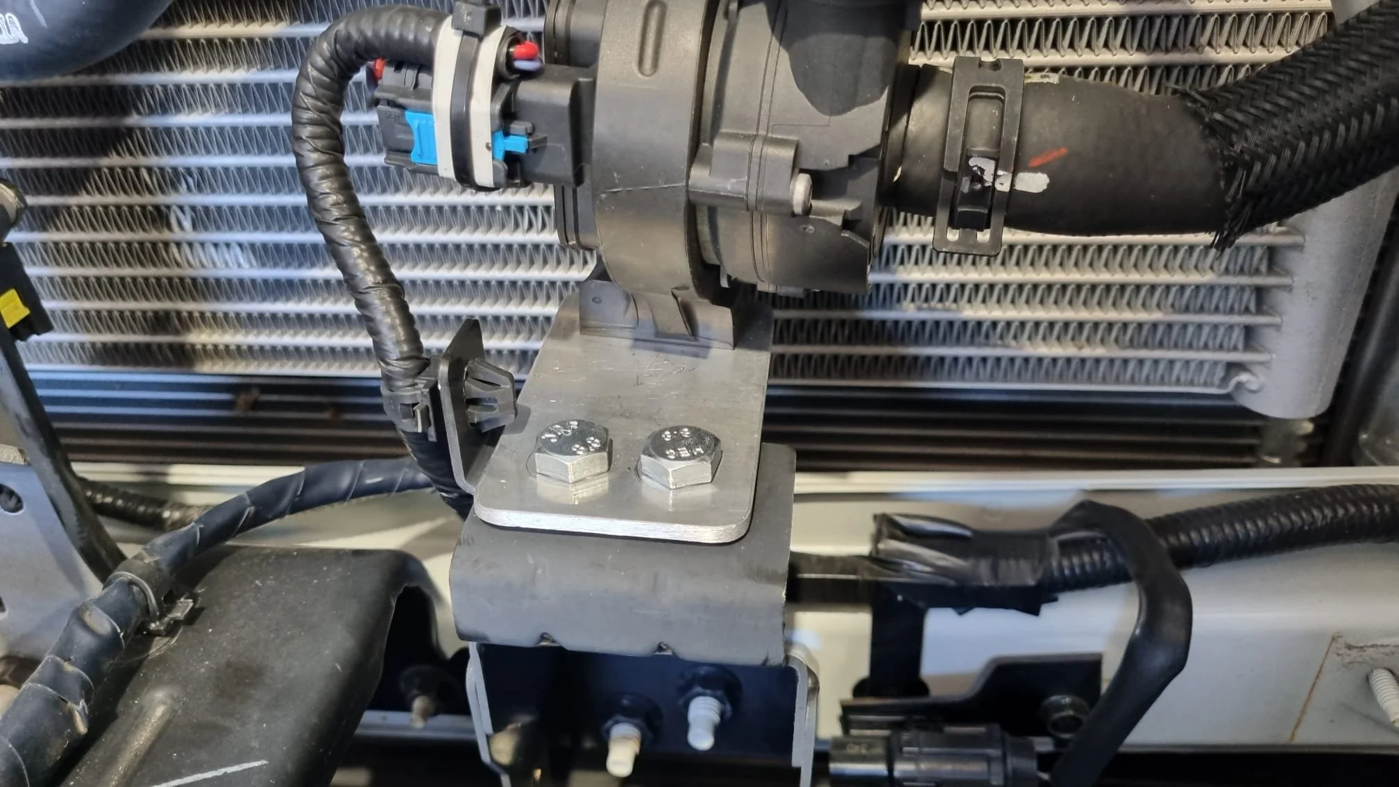 Close-up of a mechanical or electronic component mounted on a metal plate with bolts, surrounded by black cables and a cooling or heating radiator in the background.