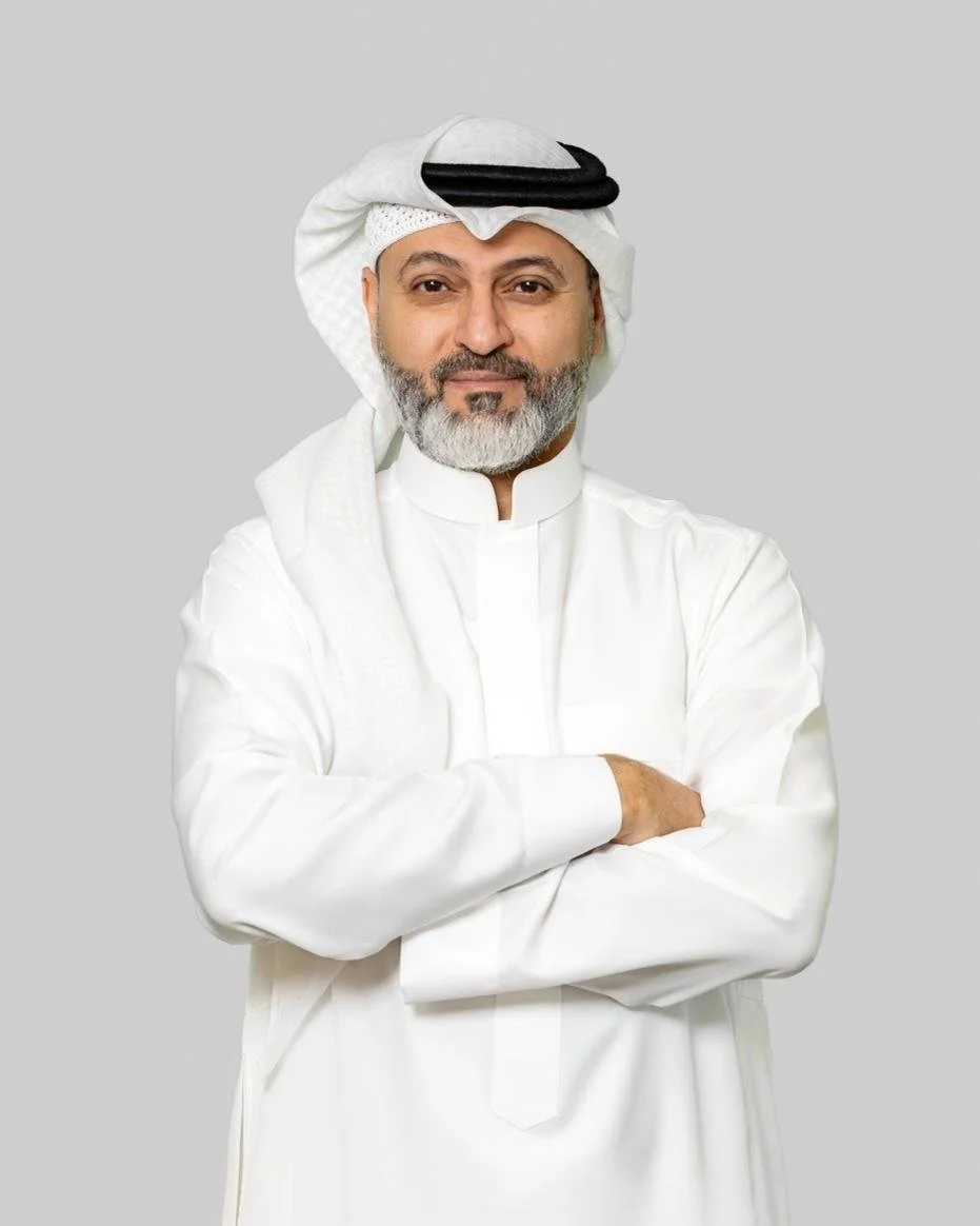 A man wearing traditional Middle Eastern attire, including a white thobe and a white headdress, standing with his arms crossed and smiling.