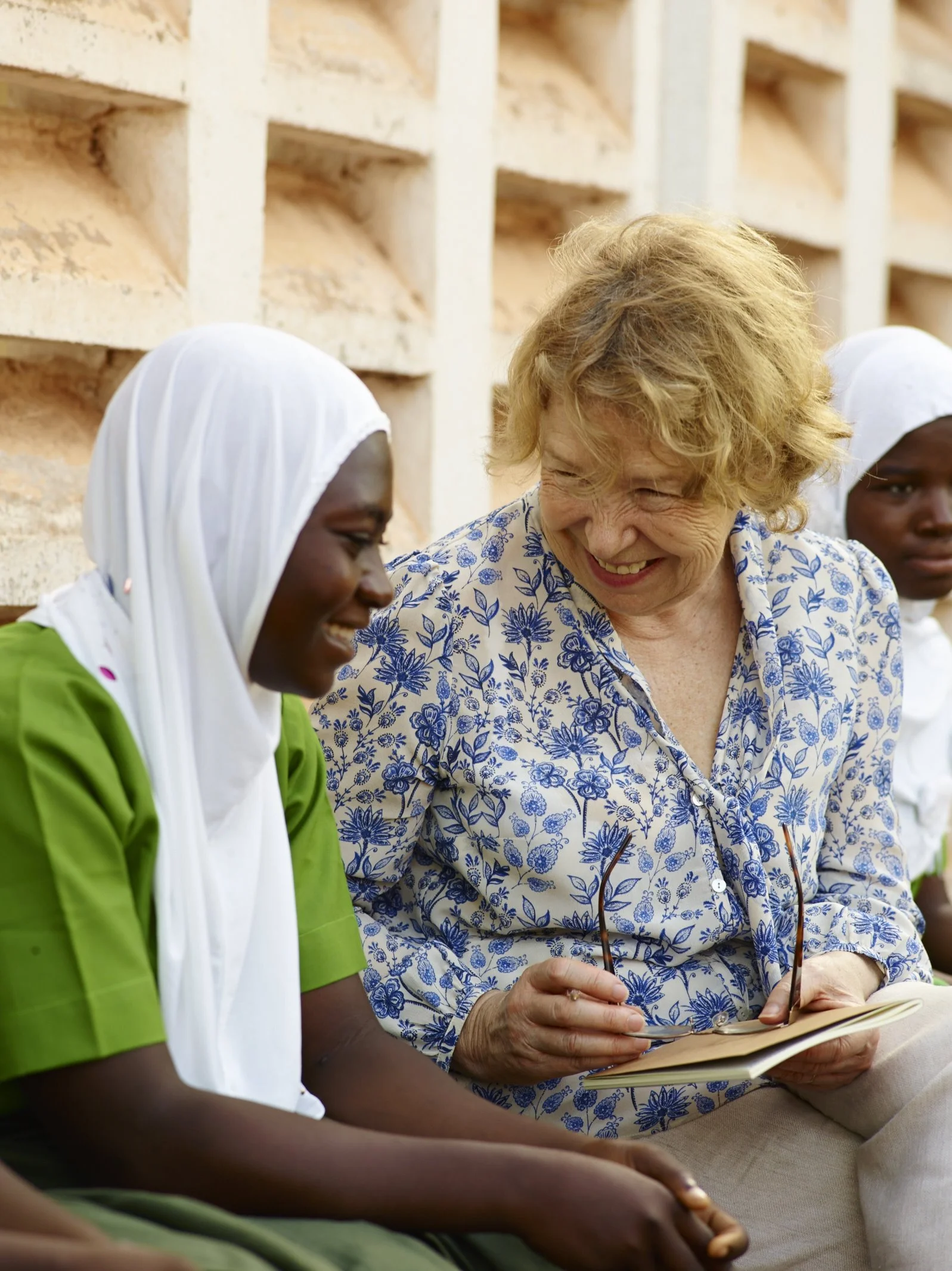 A woman with curly blonde hair smiling and holding a pair of glasses, talking with two young women wearing white headscarves and green uniforms, sitting beside a brick wall.