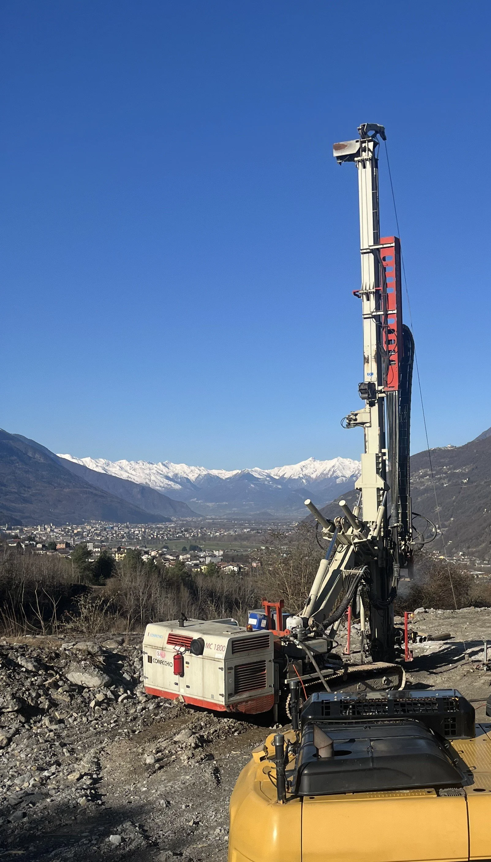 Macchina perforatrice in un sito di costruzione con montagne innevate sullo sfondo e cielo azzurro.