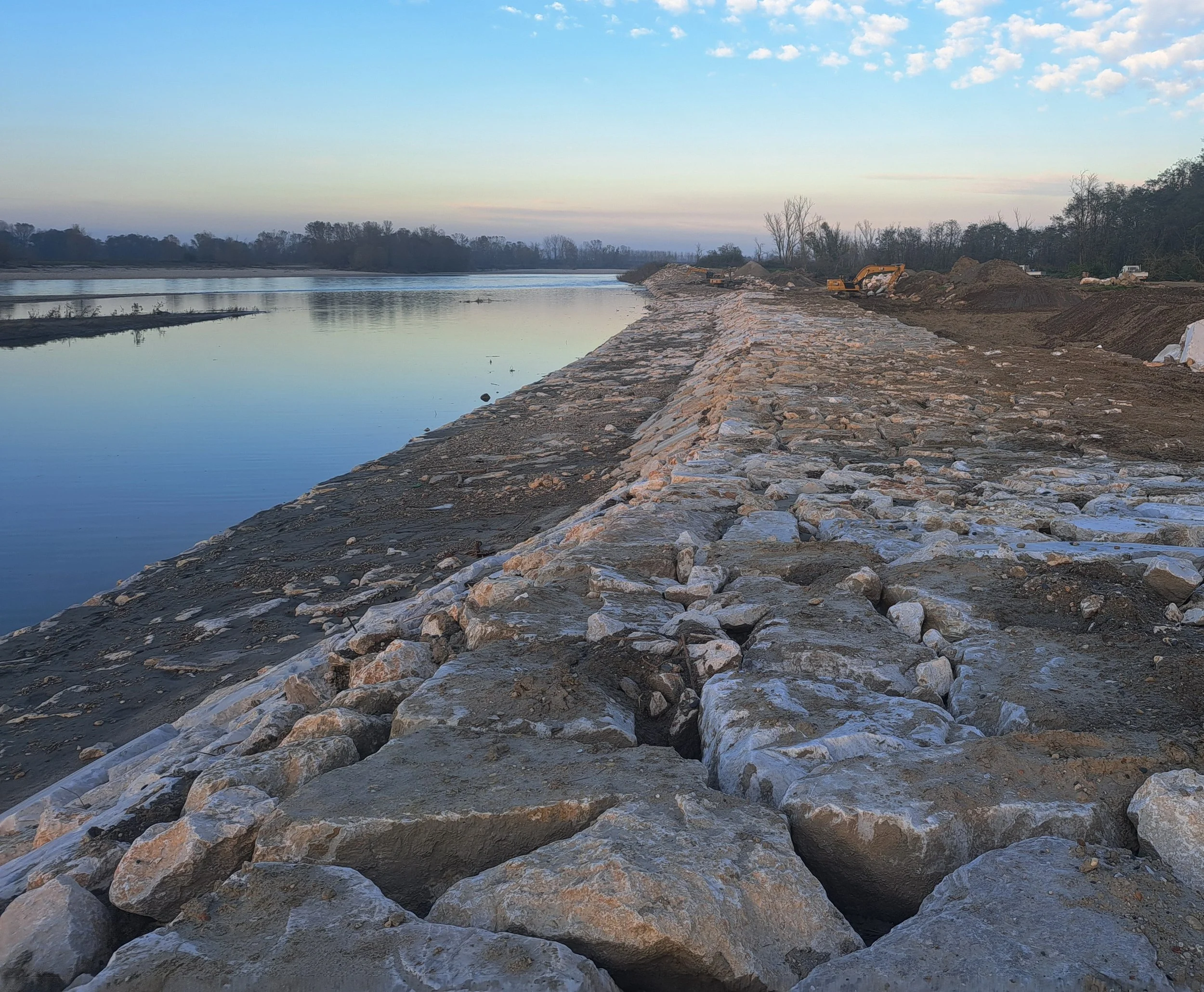 Argine di un fiume in costruzione con rocce e macchinari, cielo sereno al tramonto.
