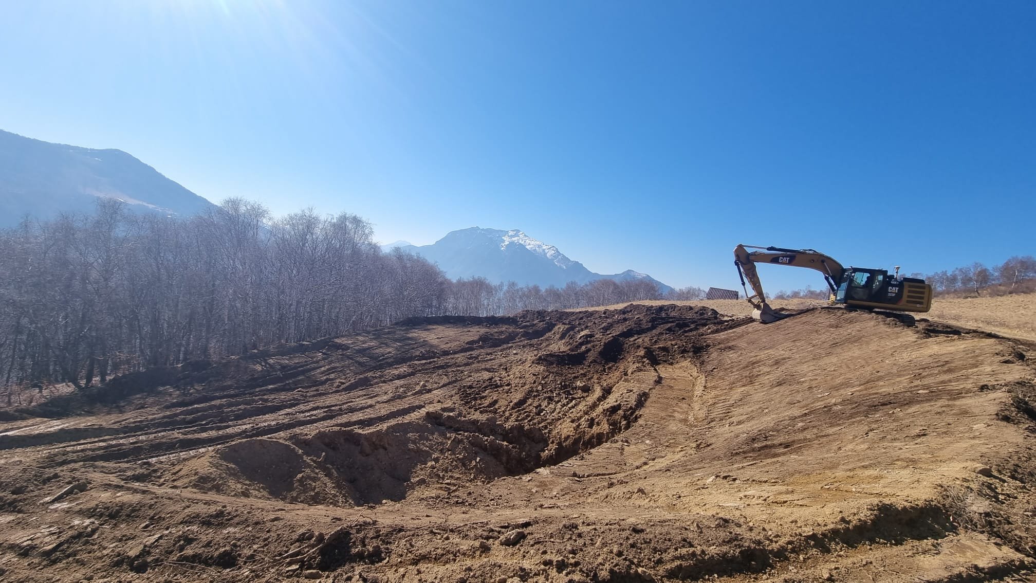 Escavatore in un cantiere su terreno collinare con montagne e alberi sullo sfondo sotto un cielo azzurro.
