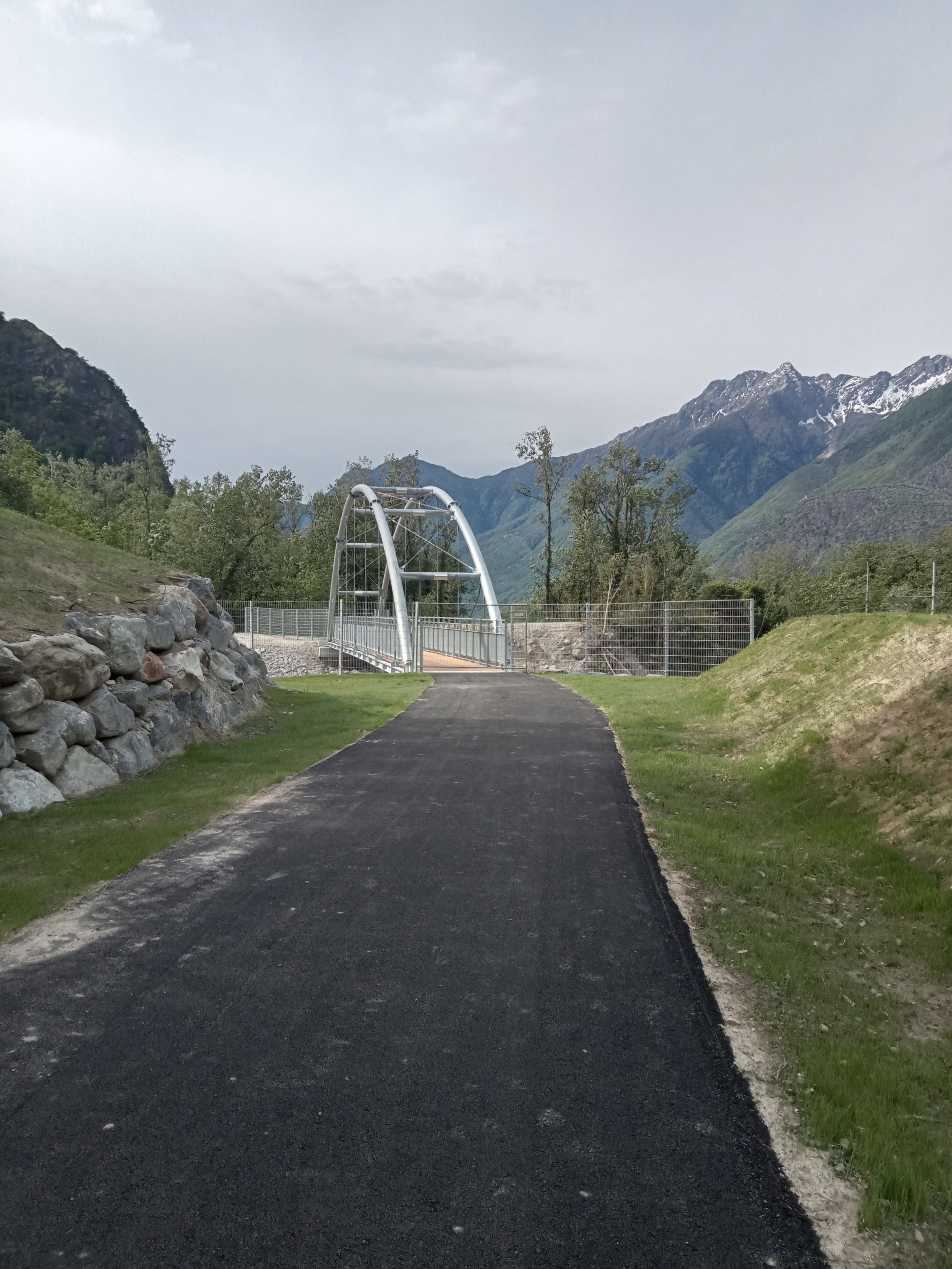 Strada asfaltata conduce a un moderno ponte in metallo circondato da colline e montagne verdi.