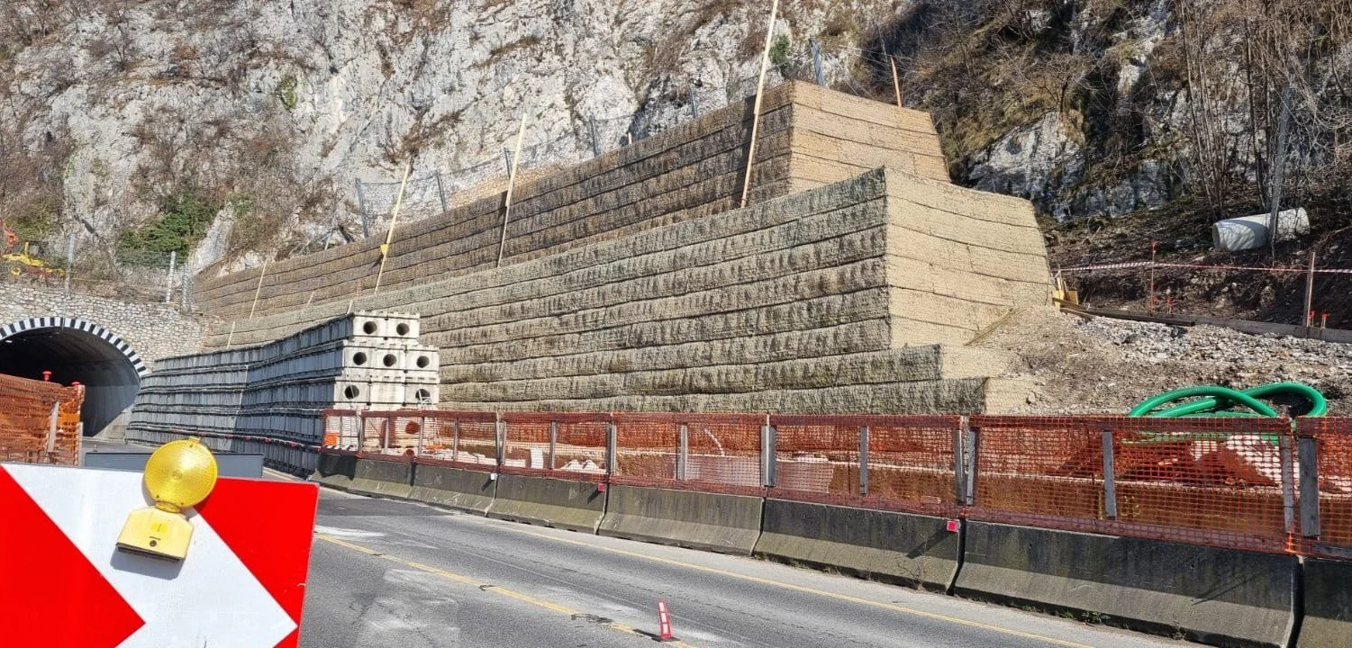 Cantieri stradali con ponteggi e materiali edili vicino a un tunnel, segnaletica stradale e montagne sullo sfondo.