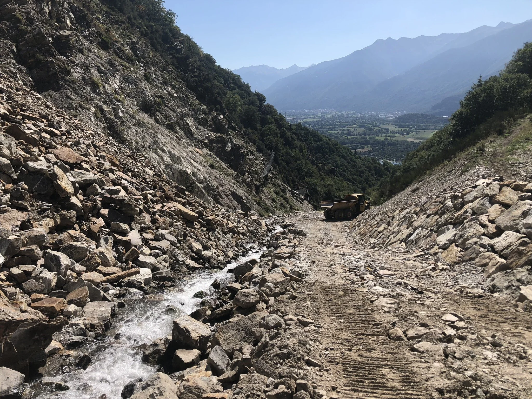 Strada sterrata in montagna con ruspa, torrente di acqua tra le rocce, vista panoramica sulla valle.