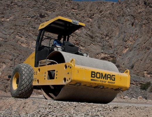 Rullo compressore giallo di marca BOMAG su terreno sterrato con sfondo montuoso.