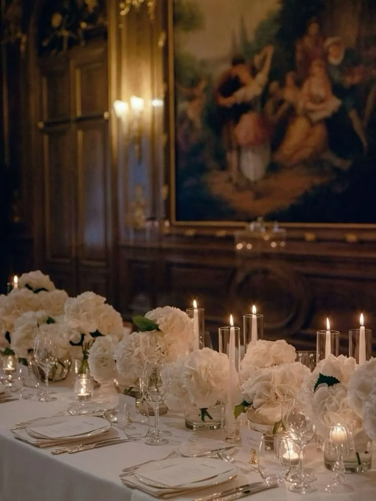 Elegant dining table decorated with white floral centerpieces and tall candles, set in a luxurious room with a large classical painting on the wall.