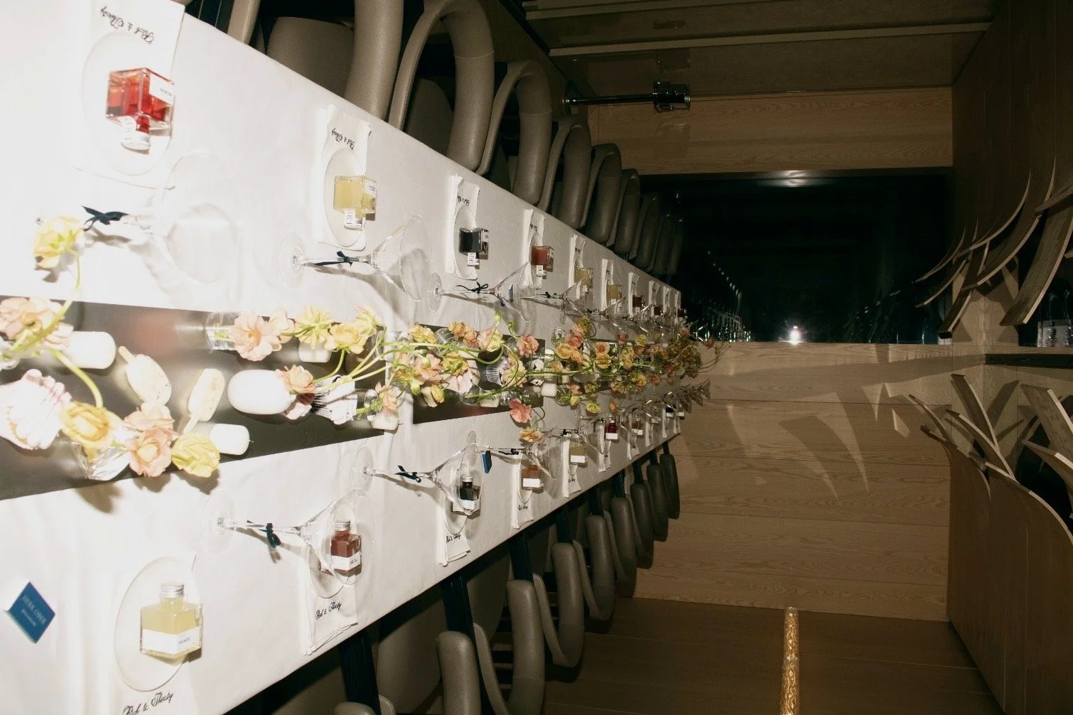 A long dining table set for a formal event with wine glasses, small bottles, and floral decorations in the center. The chairs are arranged around the table, and the setting appears elegant and prepared for guests.