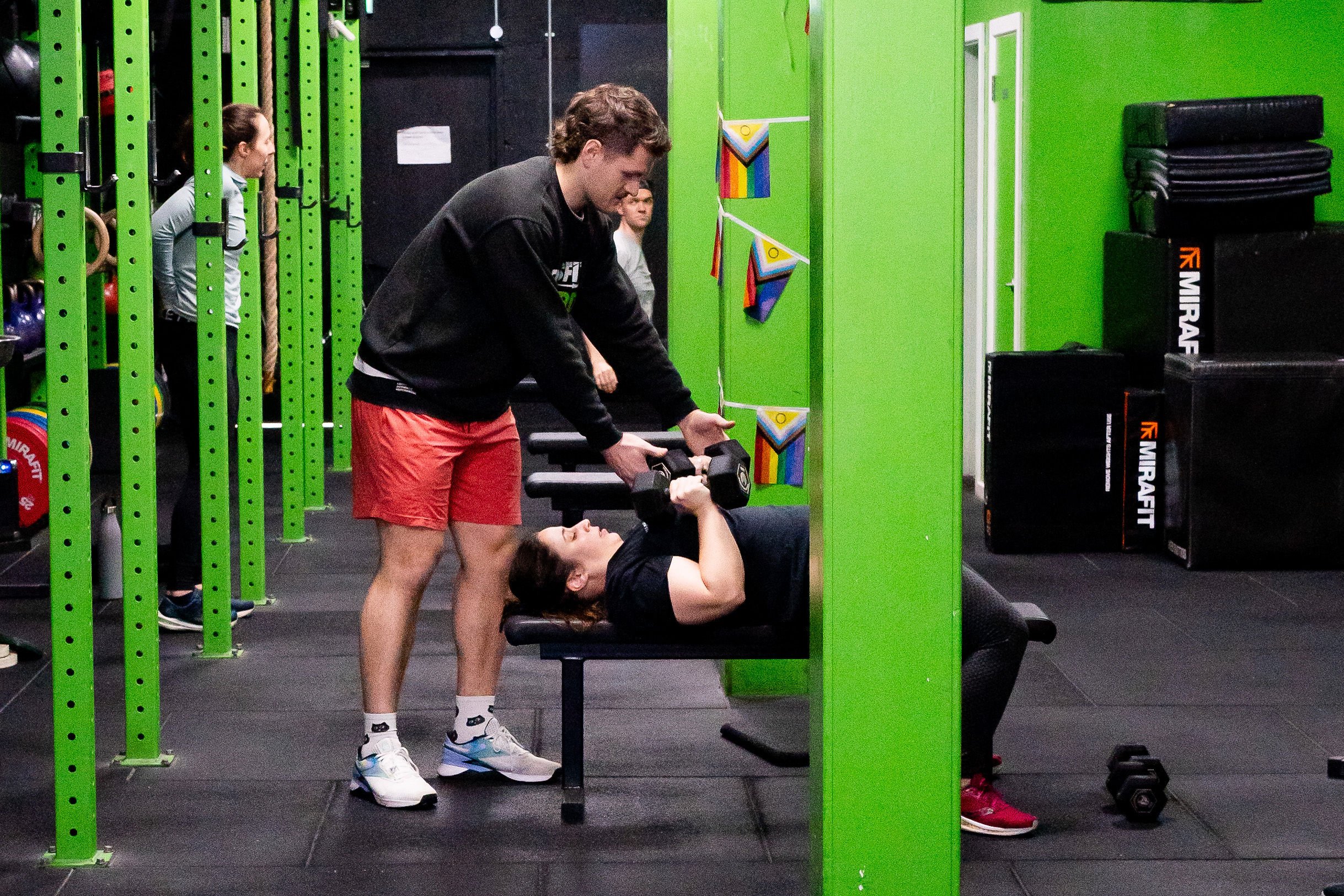 A gym scene with a person lifting dumbbells on a bench, assisted by a coach. Green workout equipment and rainbow flags are visible in the background.