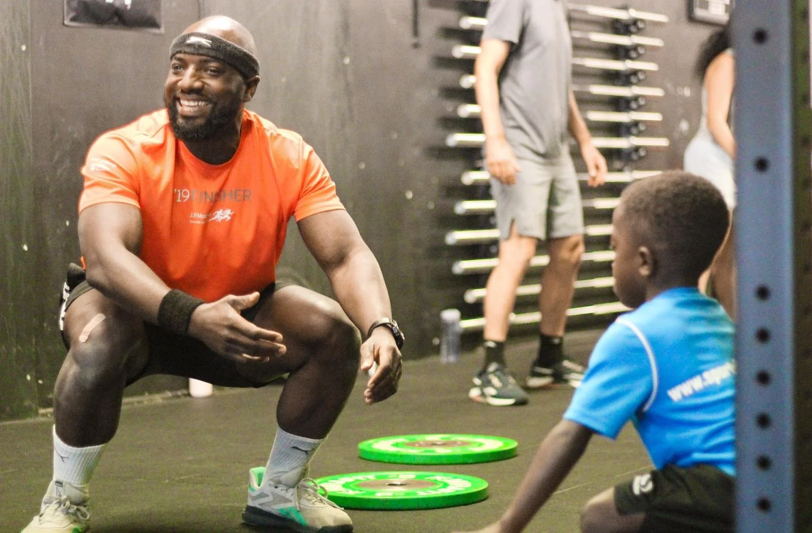 A man in an orange shirt and shorts squatting and smiling at a young boy in a blue shirt inside a gym with weights on the floor.