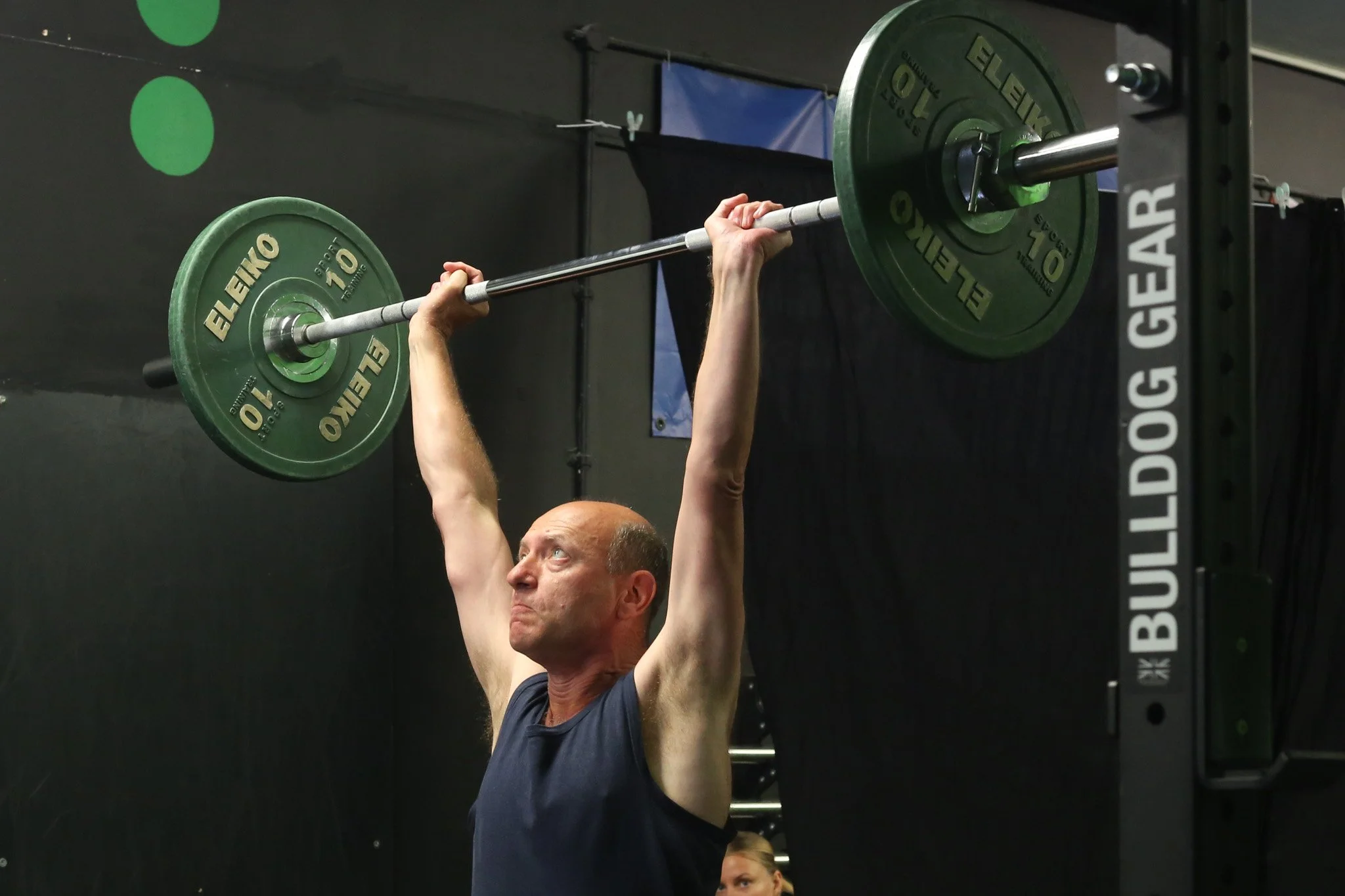 Person lifting a barbell overhead in a gym setting, with green weighted plates marked "Eleiko" and gym equipment labeled "Bulldog Gear" in the background.