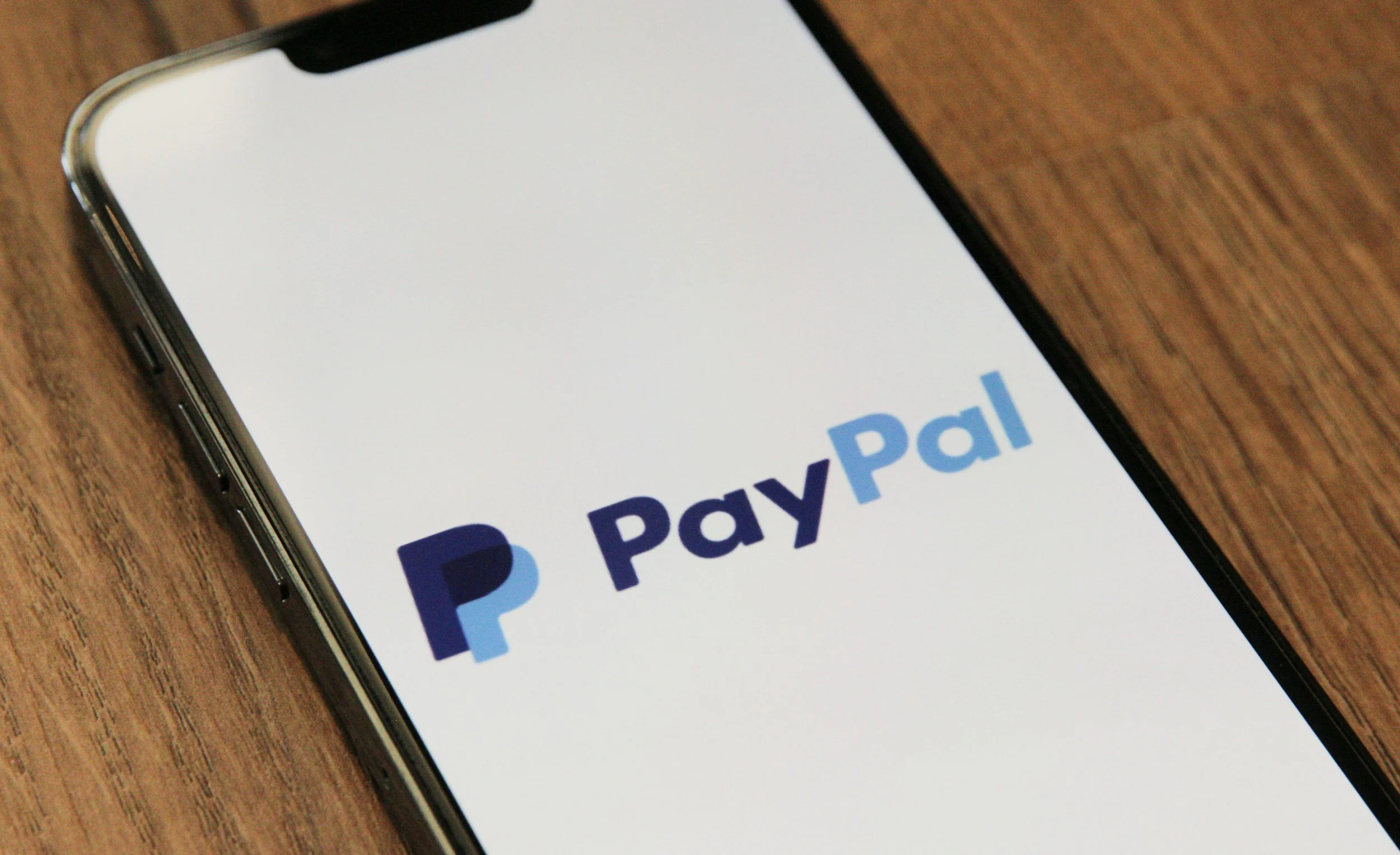Smartphone auf einem Holz Tisch zeigt die PayPal App mit Logo auf dem Bildschirm.