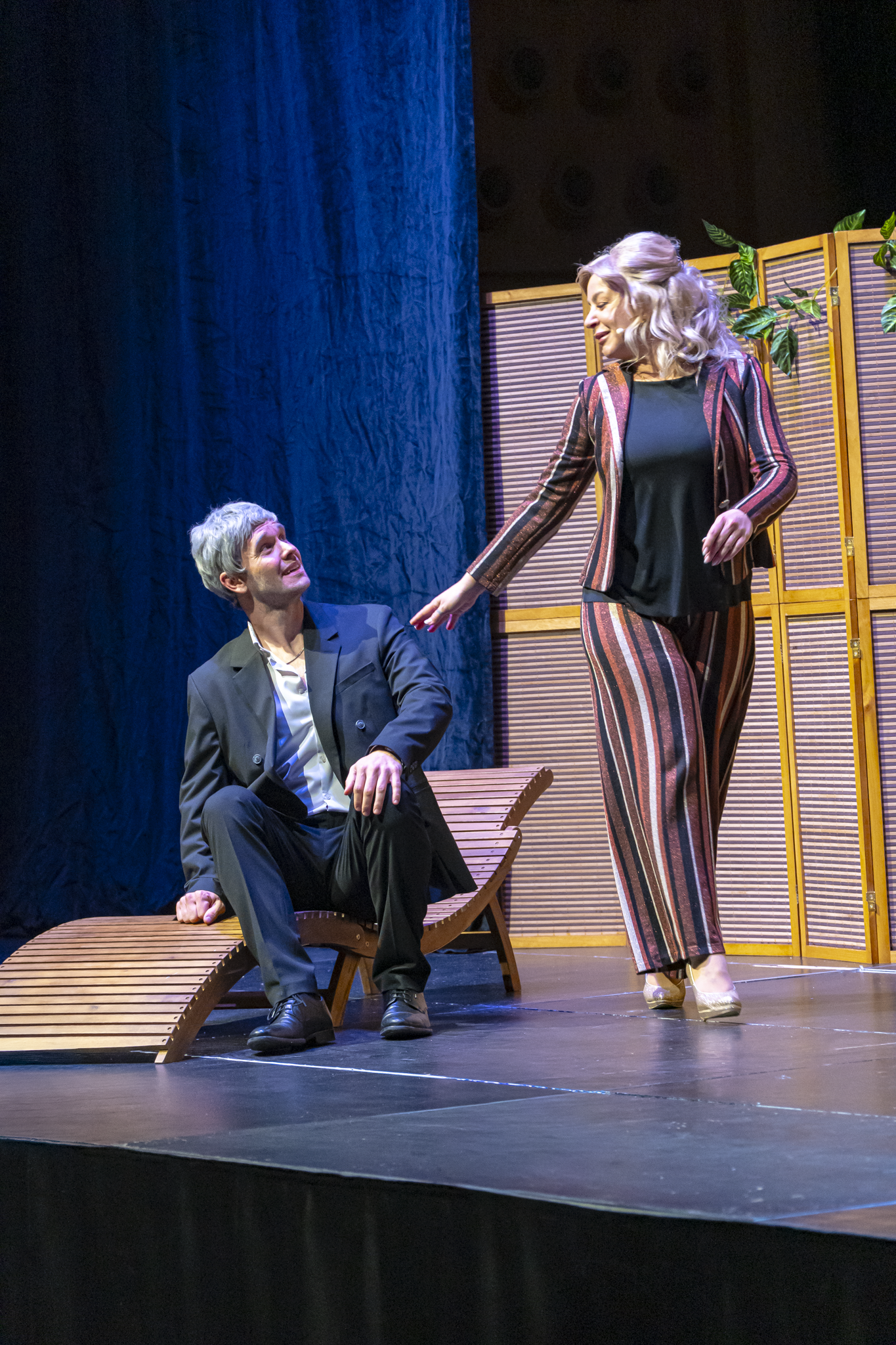 Schauspieler auf Bühne, Mann sitzt auf Liege, Frau steht neben ihm, im Theater mit Kulisse und Dekoration im Hintergrund.
