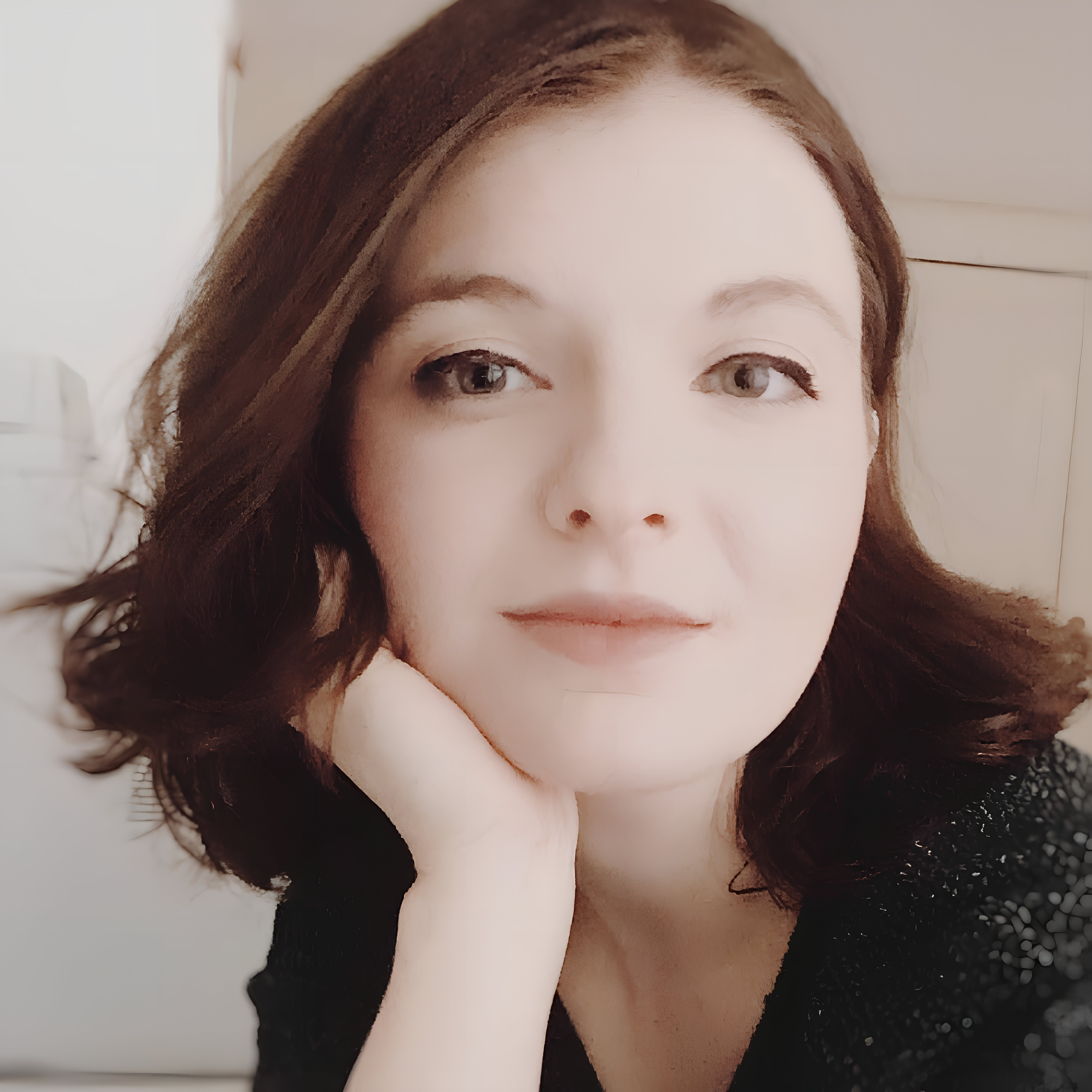 Porträt einer Frau mit braunen, gewellten Haaren, die ihre Hand an ihr Kinn legt, in einem hellen Raum.