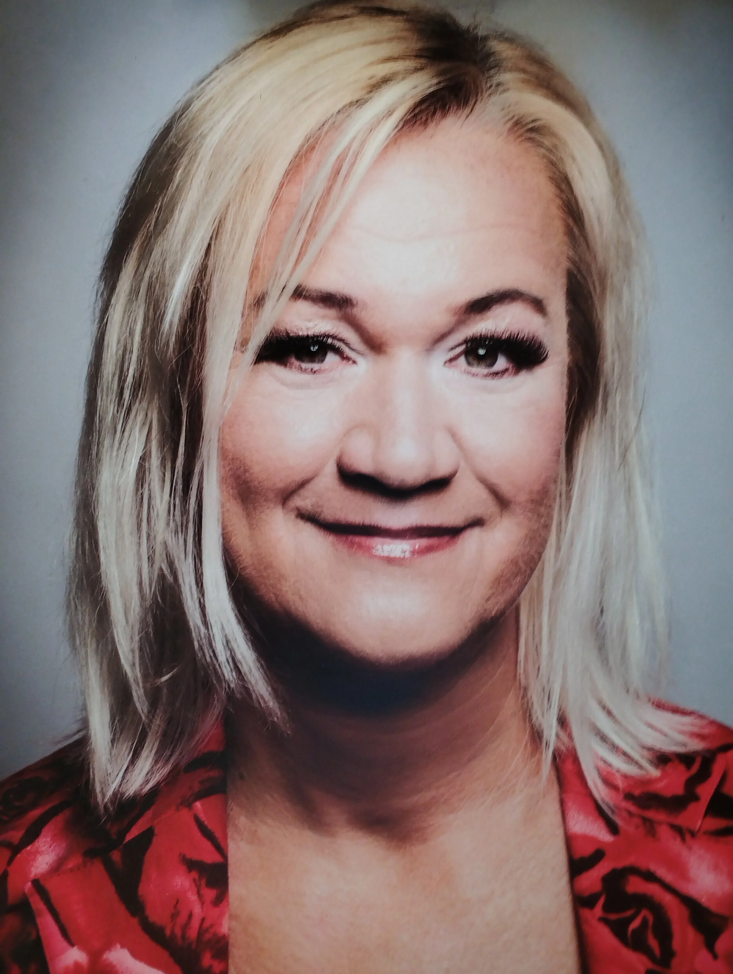 Nahaufnahme eines lachenden Frauenkopfes mit blondem, mittellangem Haar, rote Kleidung mit schwarzem Muster und natürlichem Hintergrund.