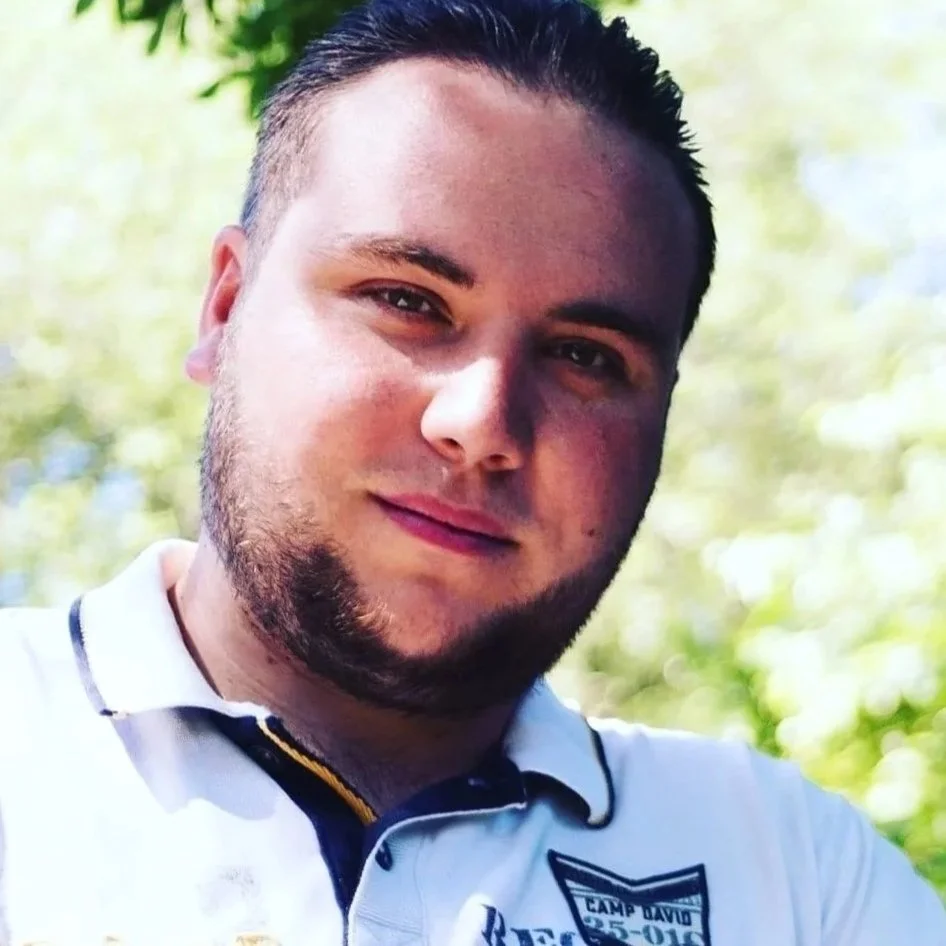 Ein junger Mann mit dunklem, kurzen Haar und Bart, trägt ein weißes Polo-Shirt mit einem Logo auf der linken Brust. Er steht im Freien vor einem Hintergrund aus grünen Blättern und Bäumen, sonniges Wetter und lächelt leicht.