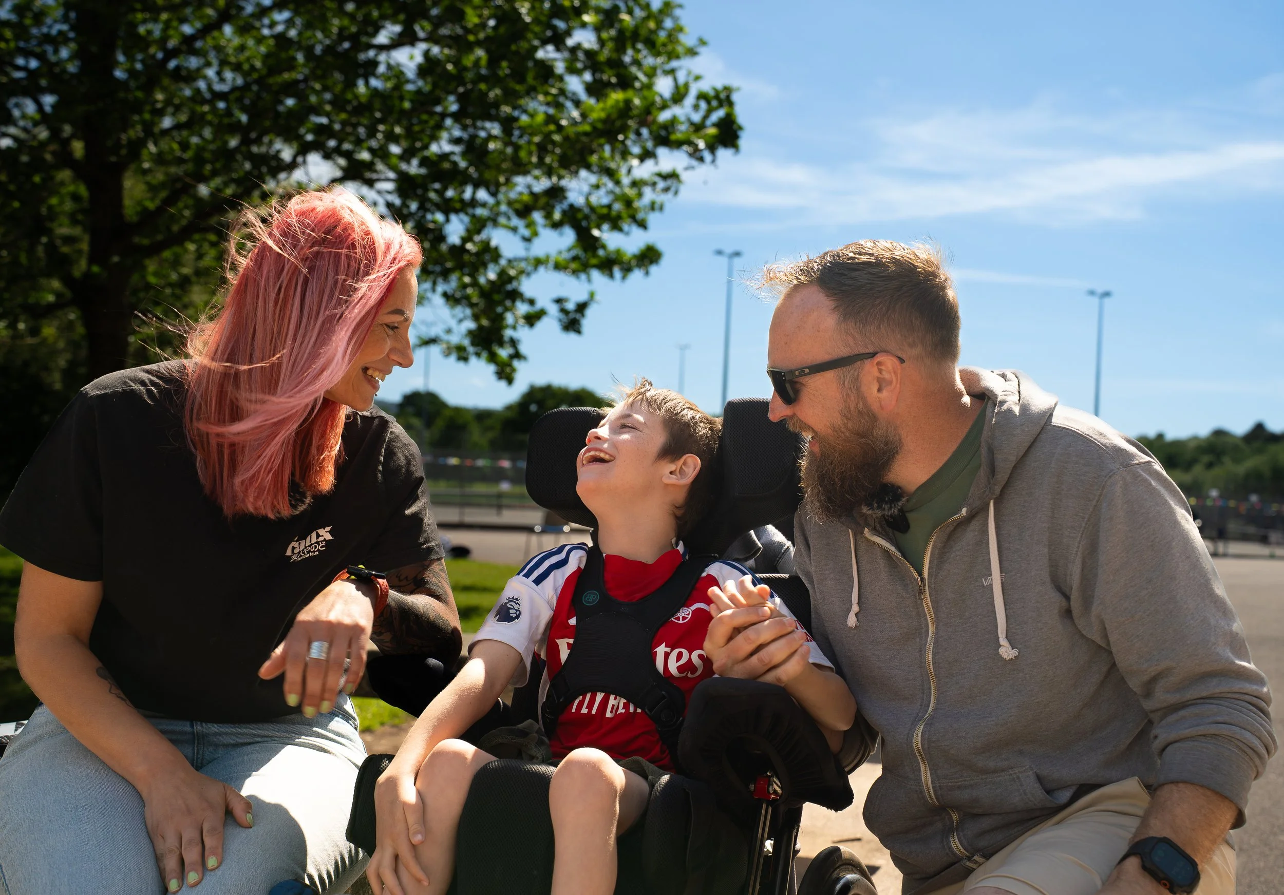 A woman with pink hair, a man with glasses, and a young boy with a disability in a wheelchair sharing a joyful moment outdoors on a sunny day.