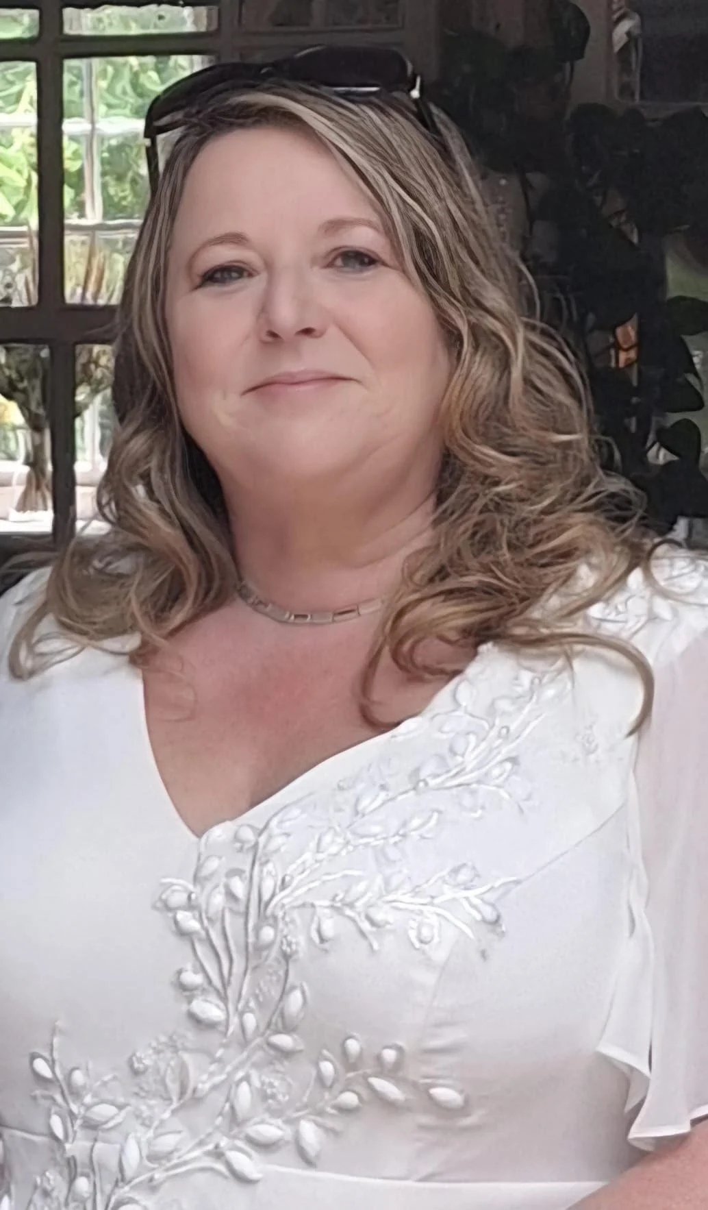 Close-up of April Griffin, administrative assistant. A woman with wavy, shoulder-length hair, wearing a white embroidered blouse, a silver necklace, and sunglasses on her head, standing indoors near a window with greenery outside.