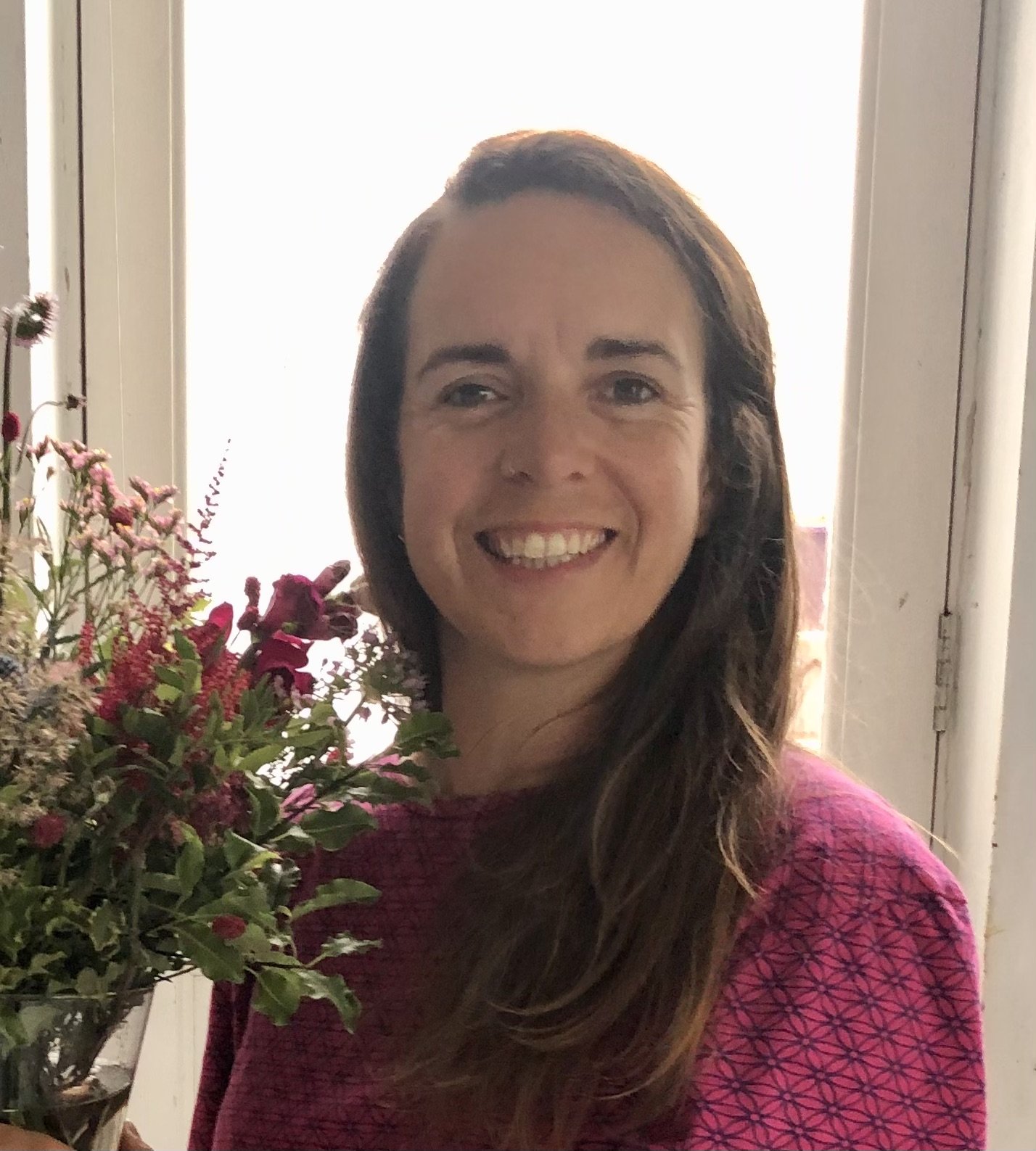 Sarah Chesney, woman with long brown hair and a bright smile standing indoors near a window, holding a bouquet of pink and purple flowers, wearing a pink shirt with a geometric pattern.