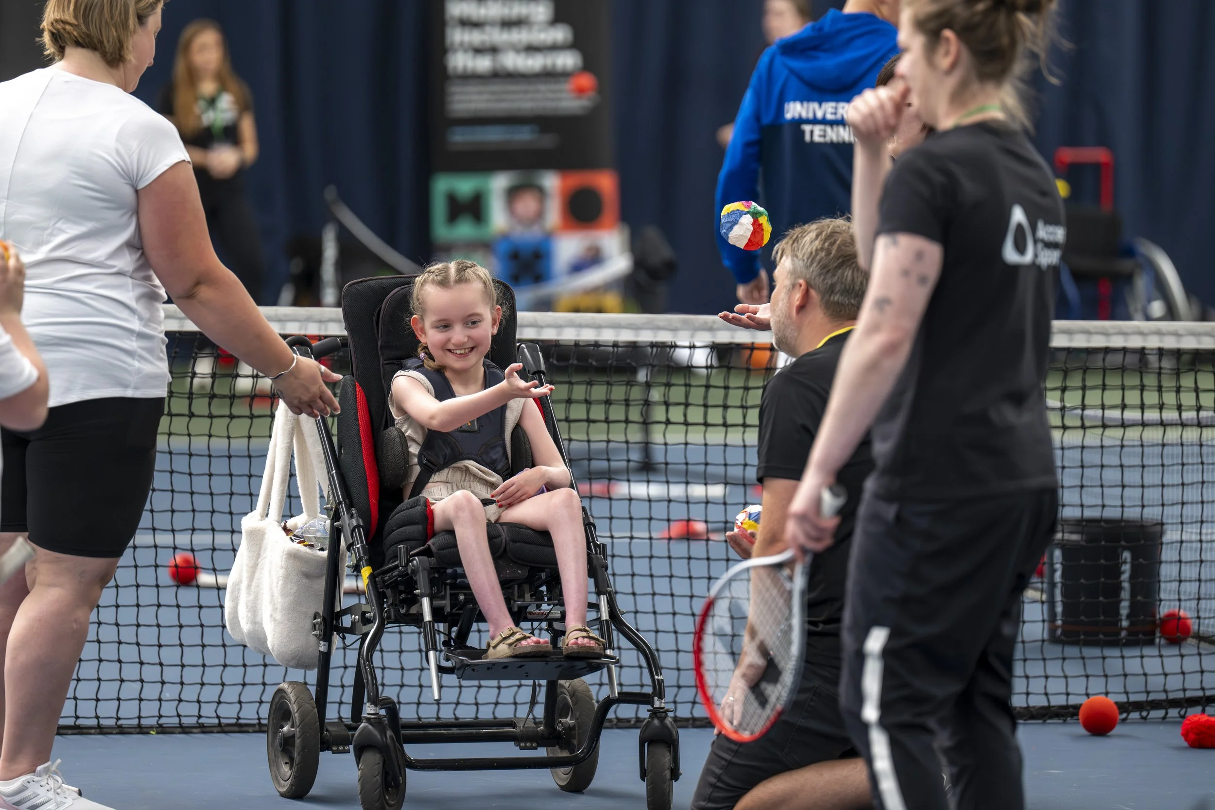 A young girl with brown hair smiling and waving from her wheelchair, surrounded by several adults on a tennis court with a net and colorful balls, with a blurred background of banners and blue curtains.