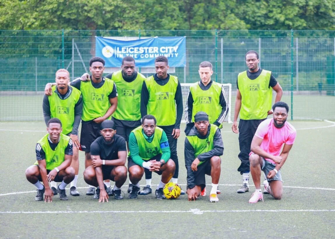 A group of ten men on a soccer field, some kneeling and some standing, wearing green and black sports gear, with a banner in the background that says "Leicester City in the Community."