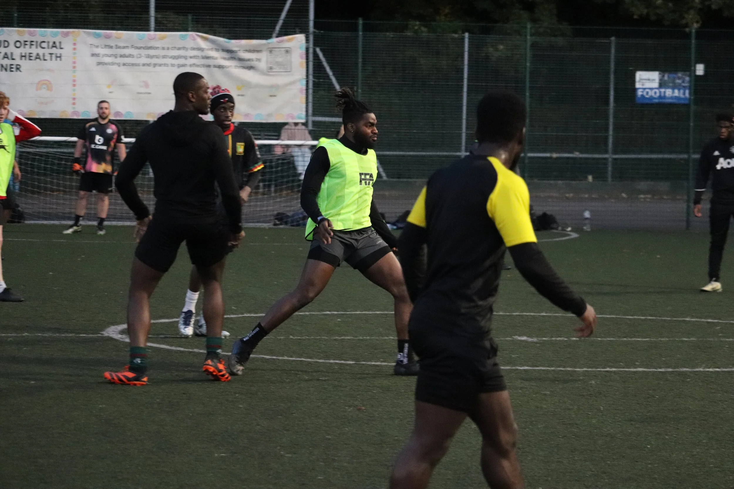 Group of athletes playing soccer on an outdoor field, some wearing black and yellow jerseys, others in practice vests, with a goal and fence in the background.