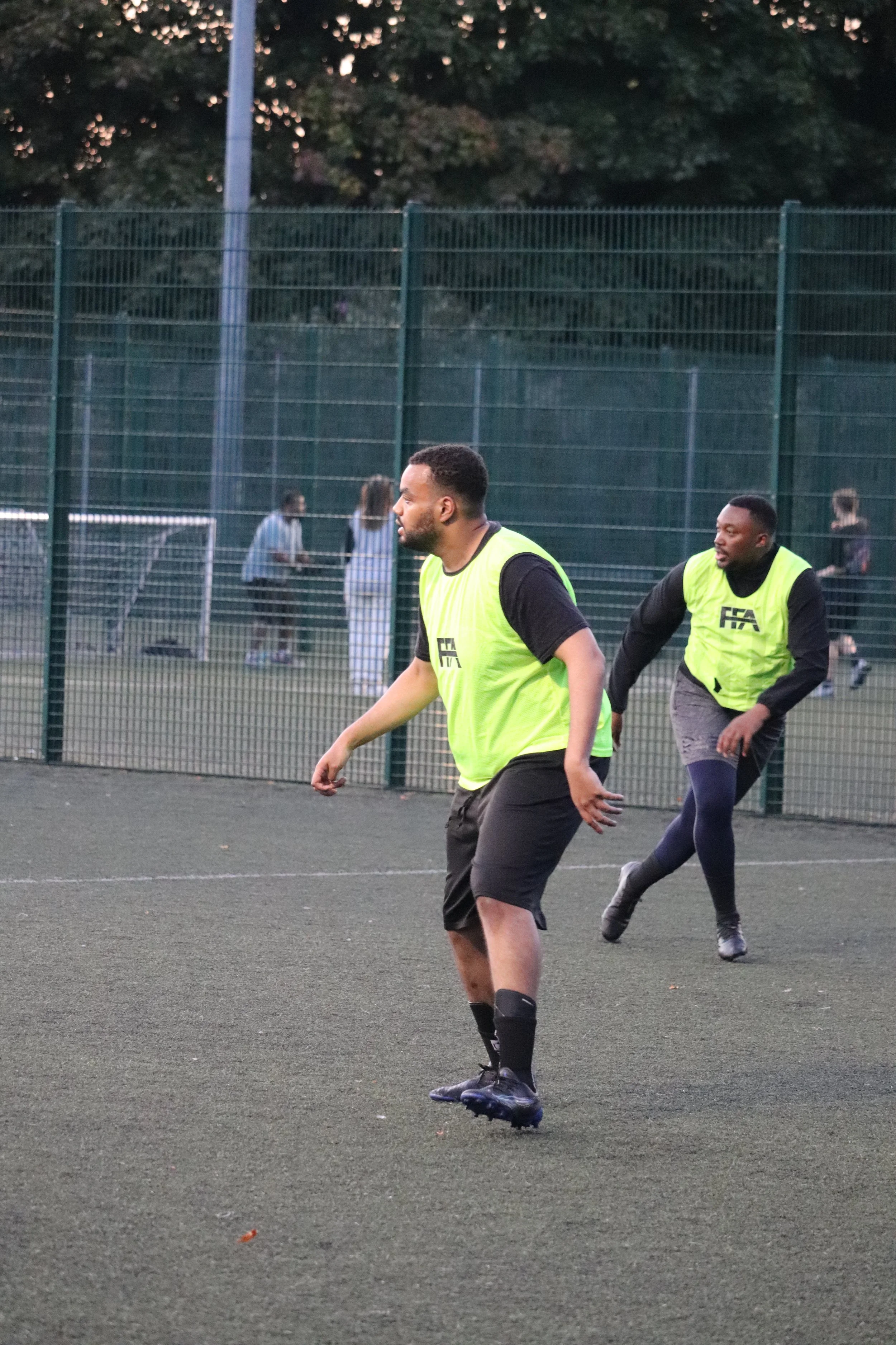 Two men playing soccer on an outdoor field, wearing yellow practice vests and black athletic clothing.