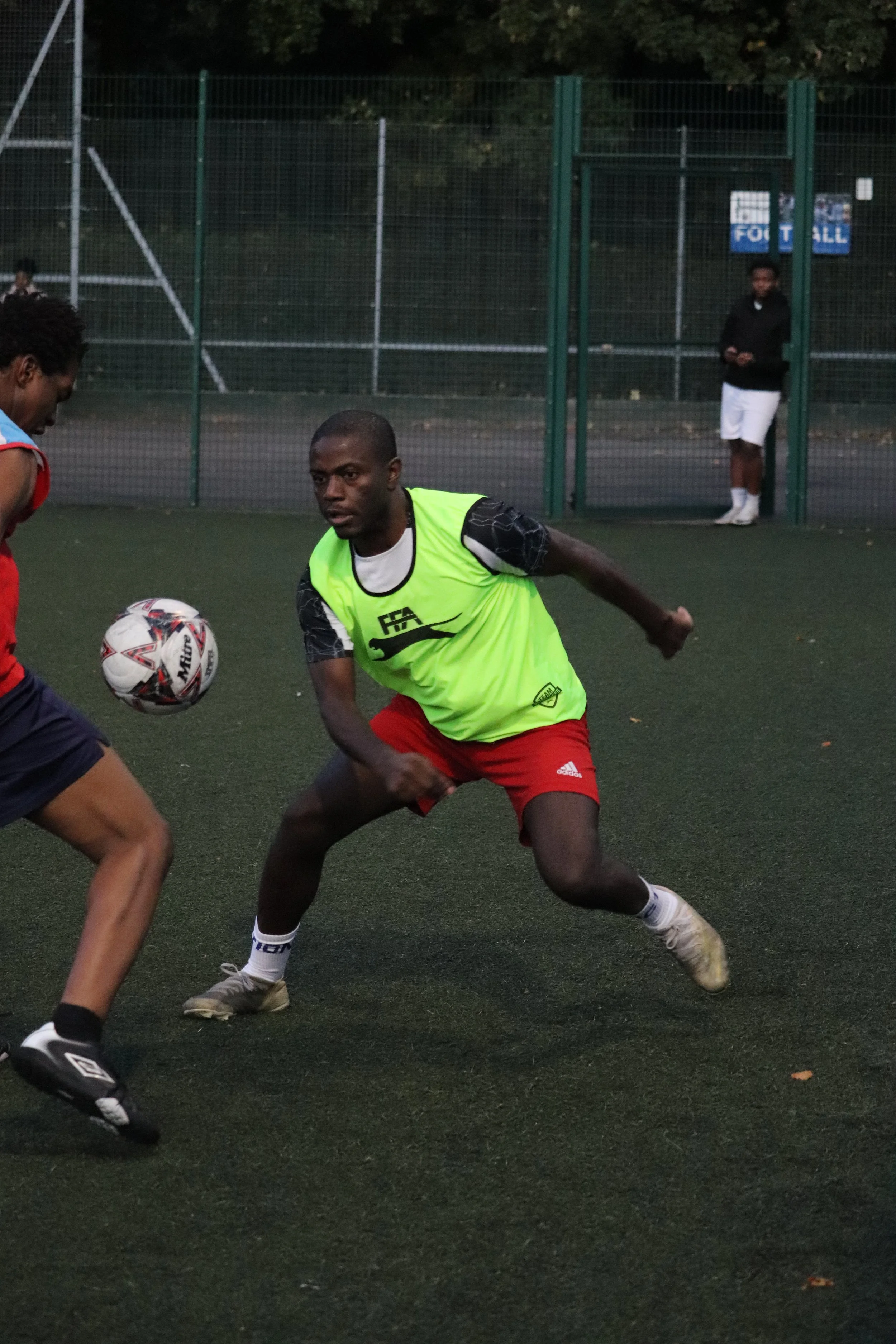 Two soccer players competing for control of a soccer ball on an outdoor field, with a person standing near a fence in the background.