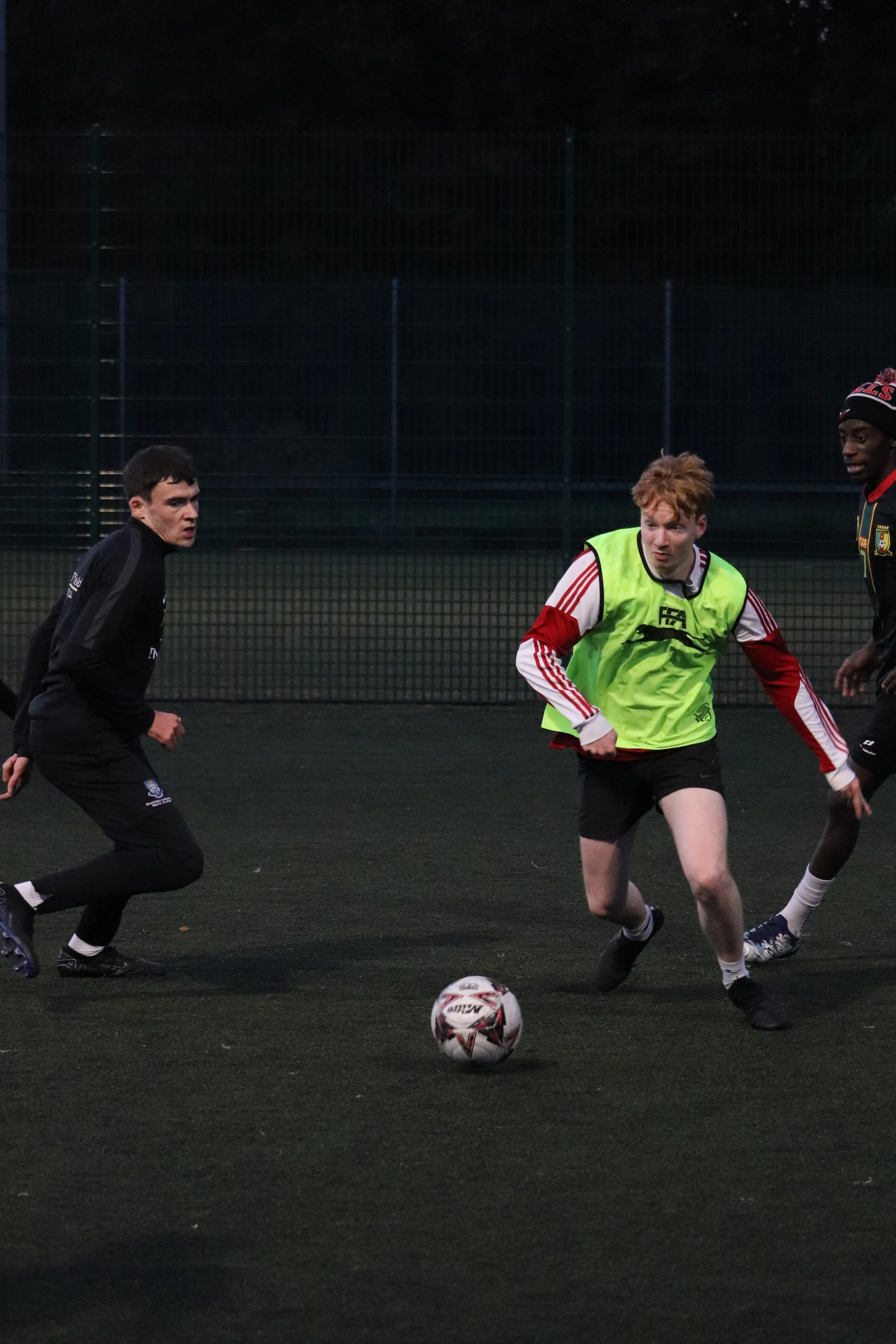 Three young men playing soccer on an outdoor field during evening, one wearing a bright yellow vest, another in a dark jacket, and the third in a black shirt and shorts, with a soccer ball on the ground.