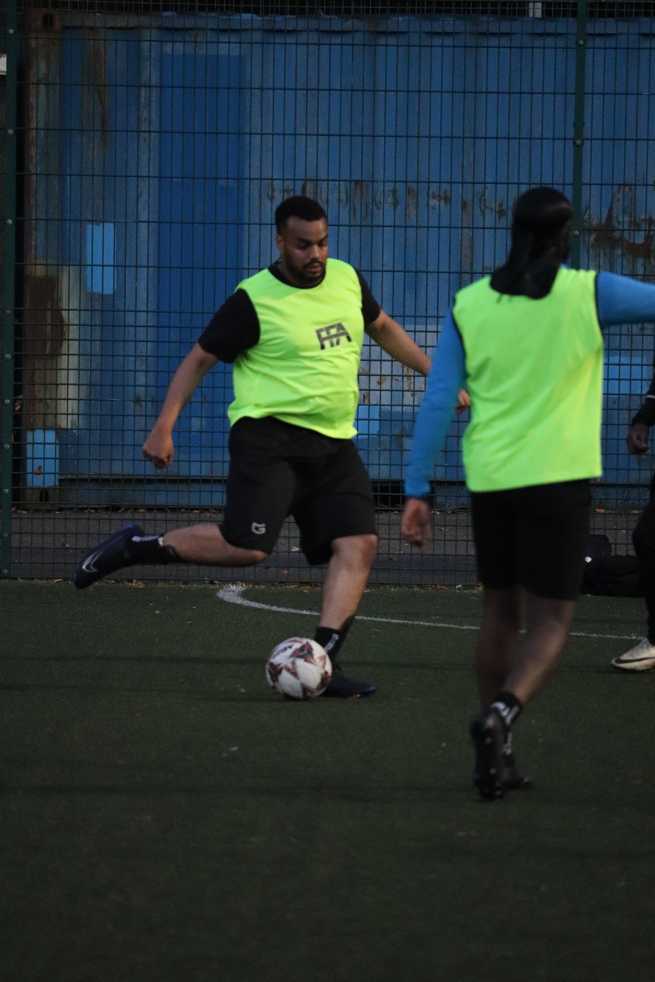 Two players in fluorescent green vests playing soccer on a turf field, one about to kick the ball.