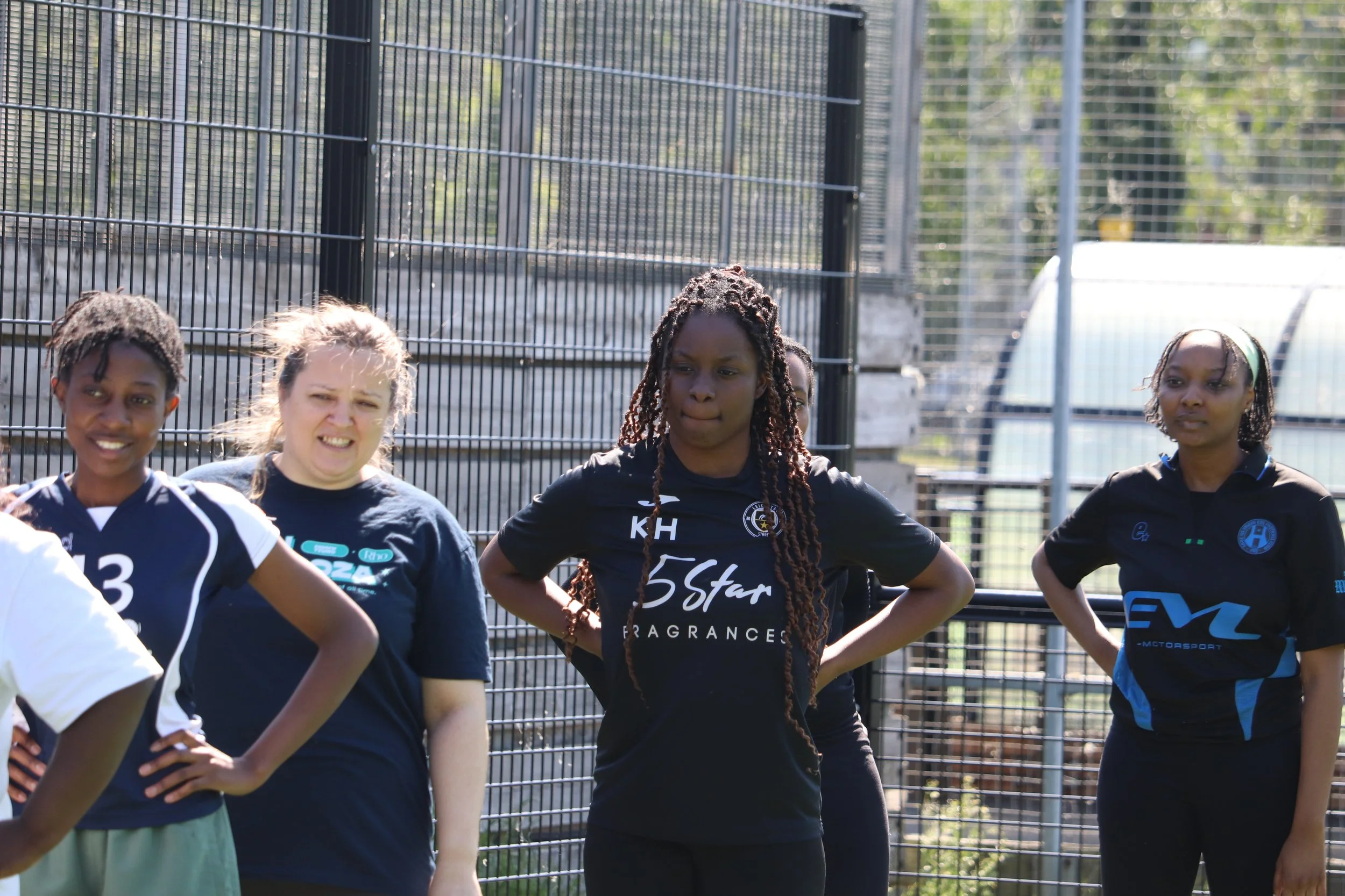 Group of women standing in front of a wire fence during daytime, some with their hands on hips, wearing sports or team uniforms.