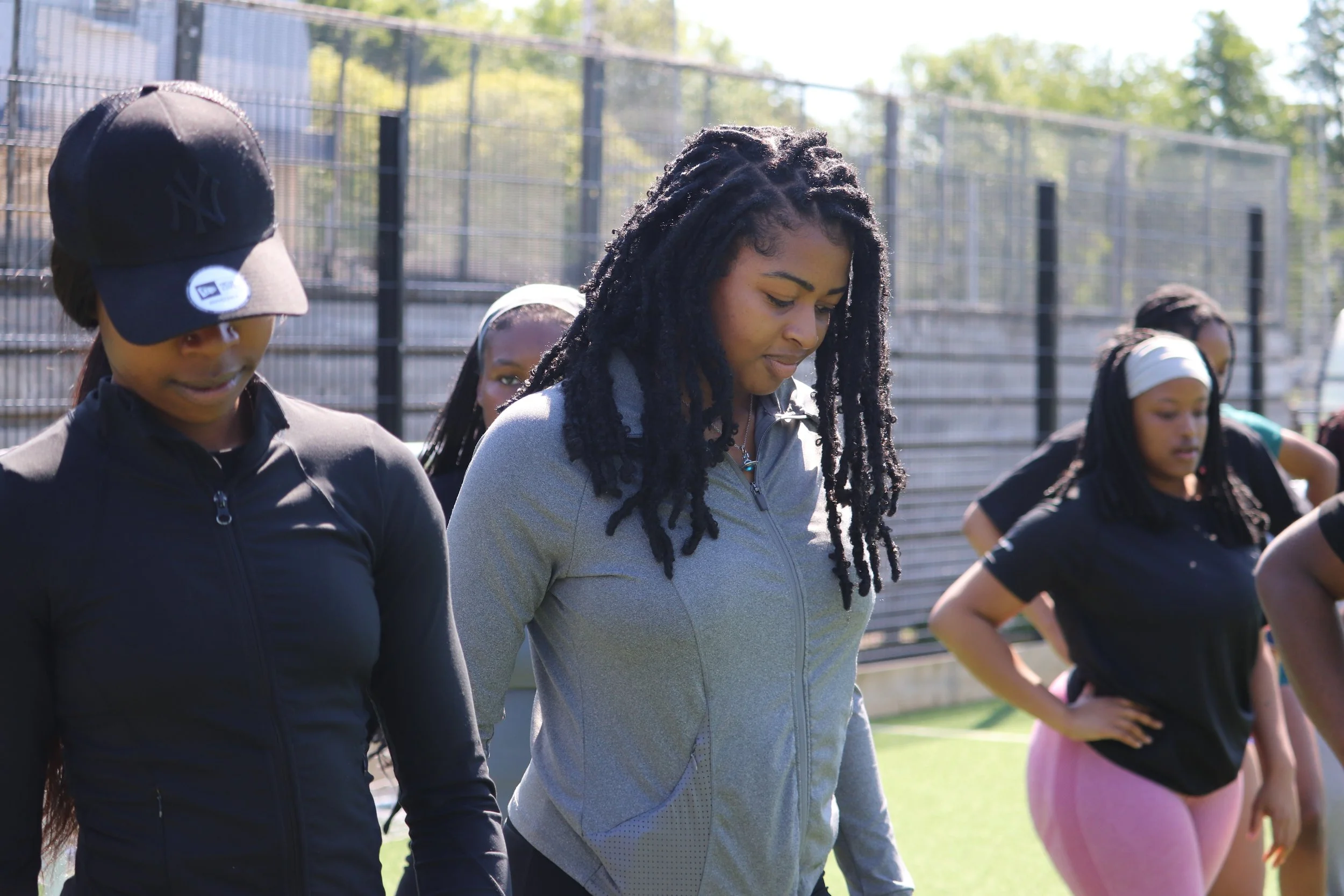 Women standing outdoors on a grassy field with a high fence in the background, possibly during a workout or training session.