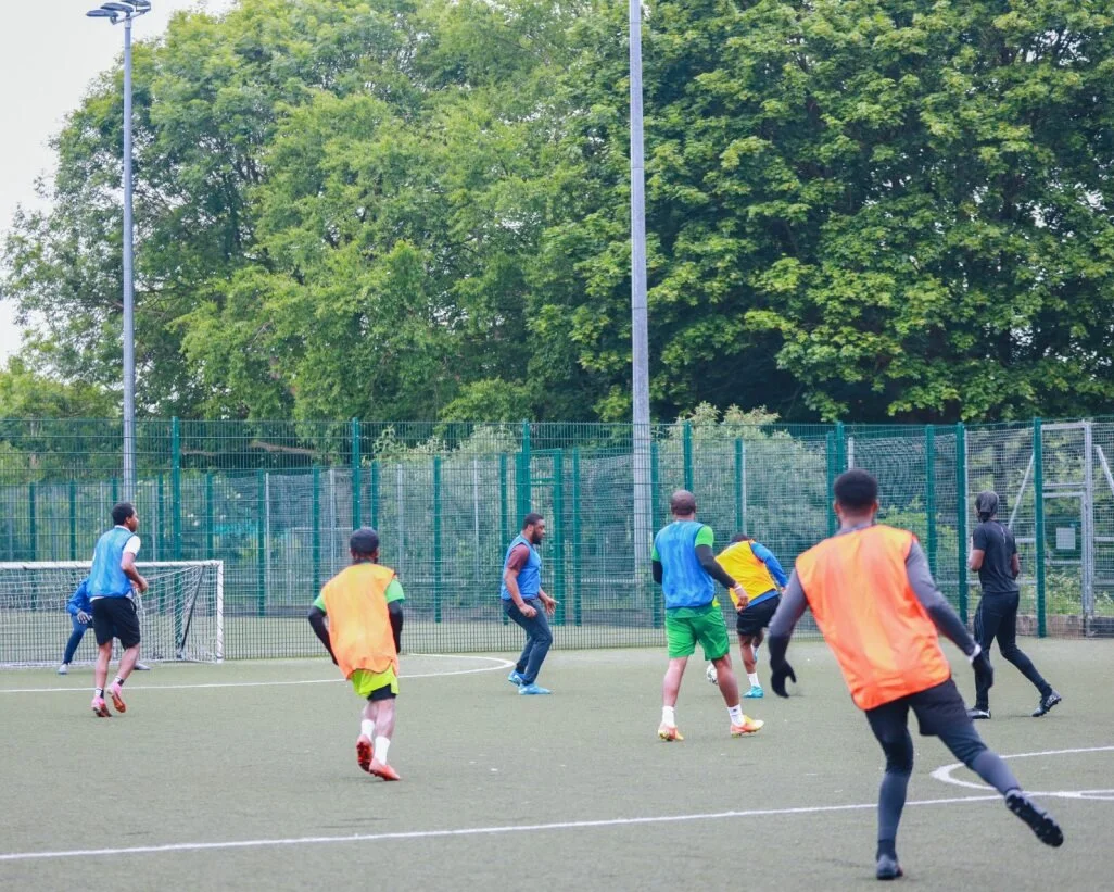 Soccer players practicing on an outdoor field, some wearing blue and orange training vests, with a goal post and tall green trees in the background.