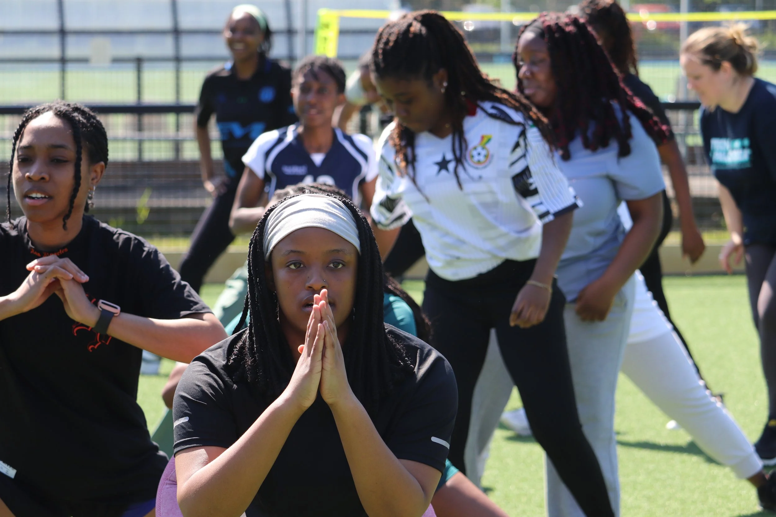 A group of women and girls participating in an outdoor group exercise or stretching session on a field, with some women in athletic attire and others in casual clothing.