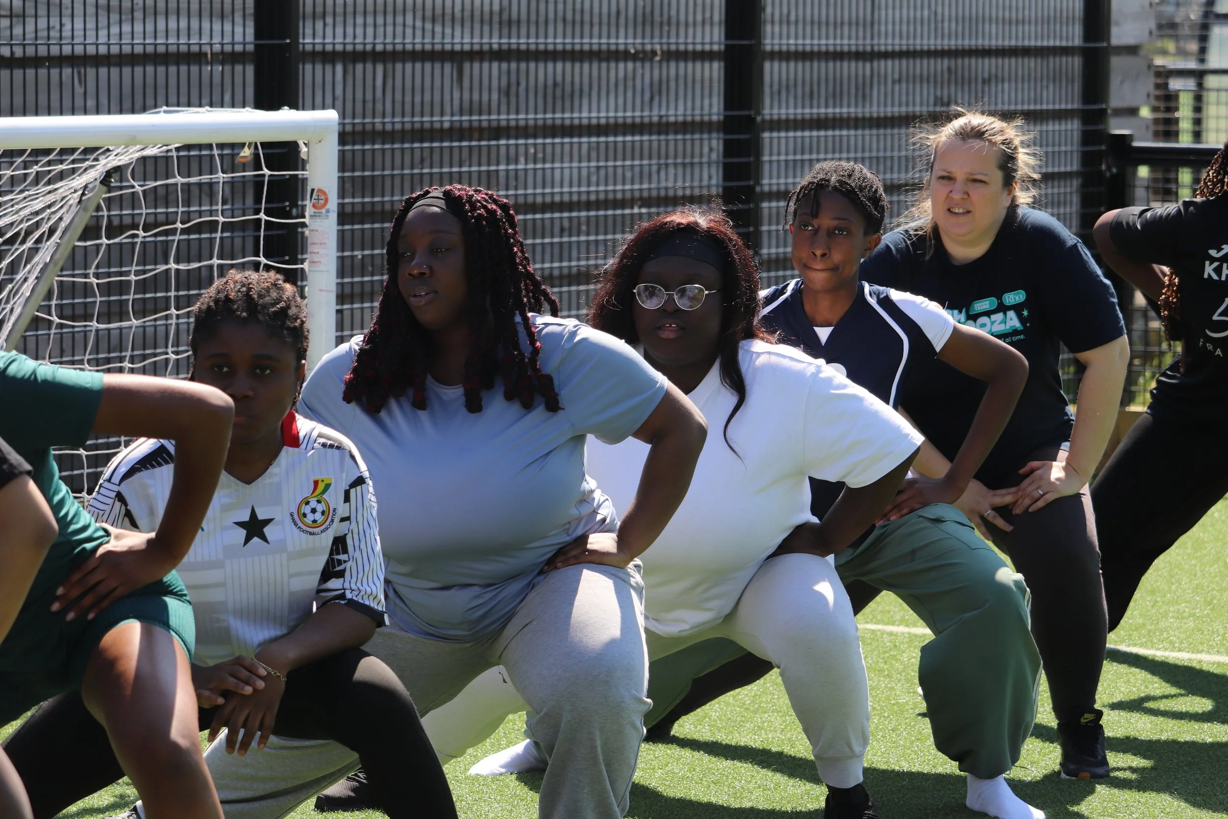 Women and young girls participating in a group activity on a soccer field, standing in a ready stance, near a goalpost with a fence in the background.