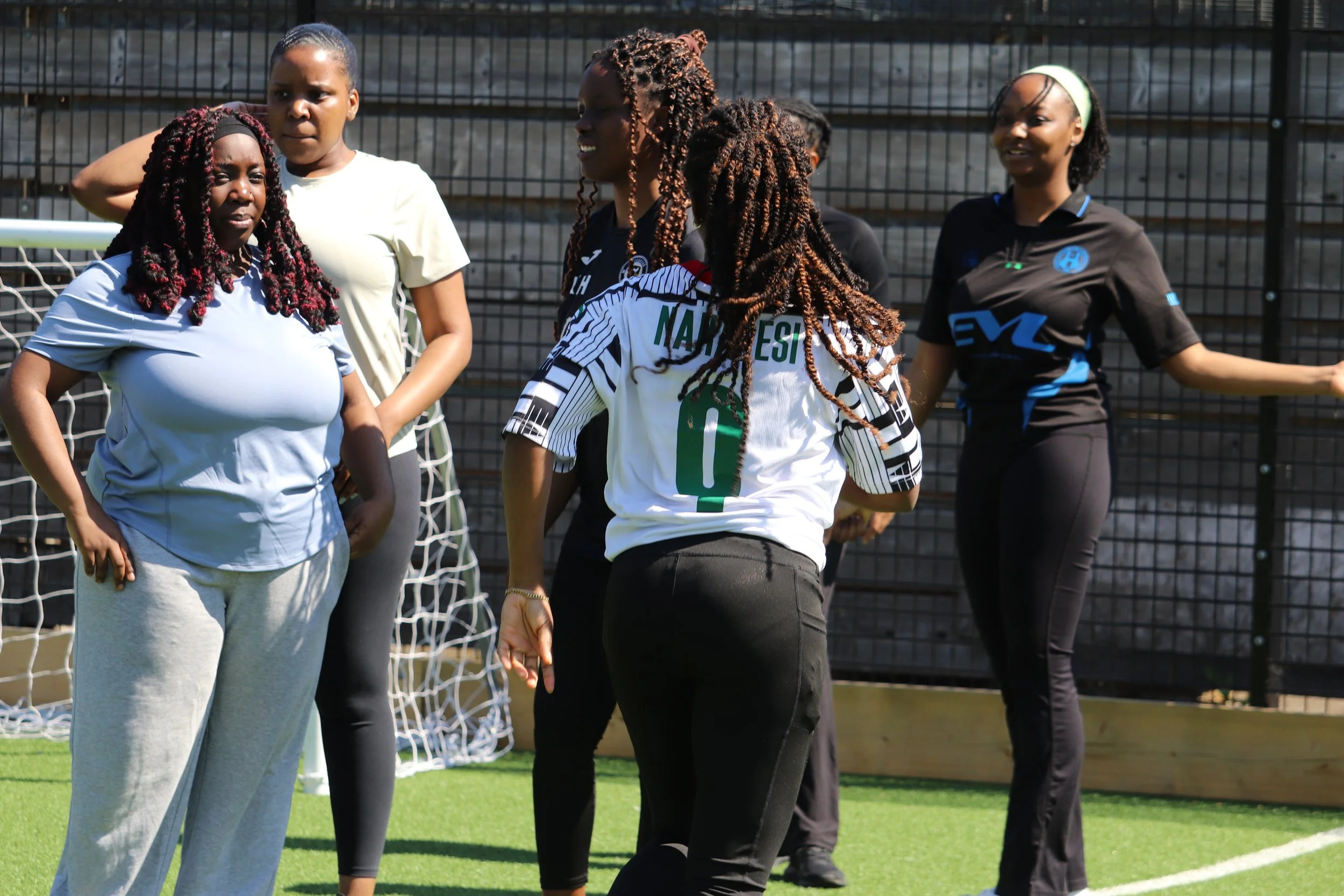Group of women on a soccer field near a goalpost, engaged in conversation, with a black fence in the background.