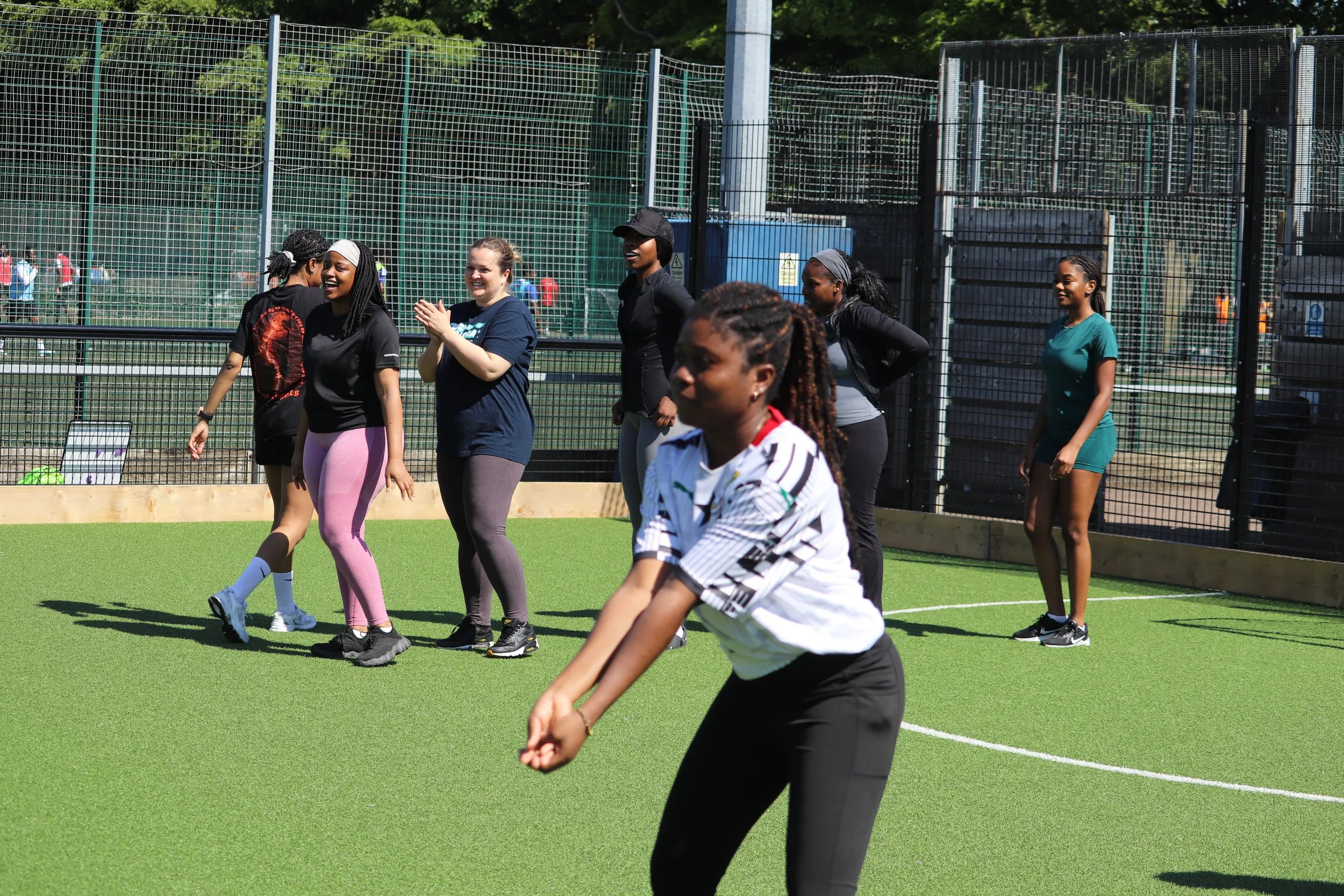 Group of women and girls playing or engaging in an activity on a green outdoor sports field surrounded by a tall safety fence.