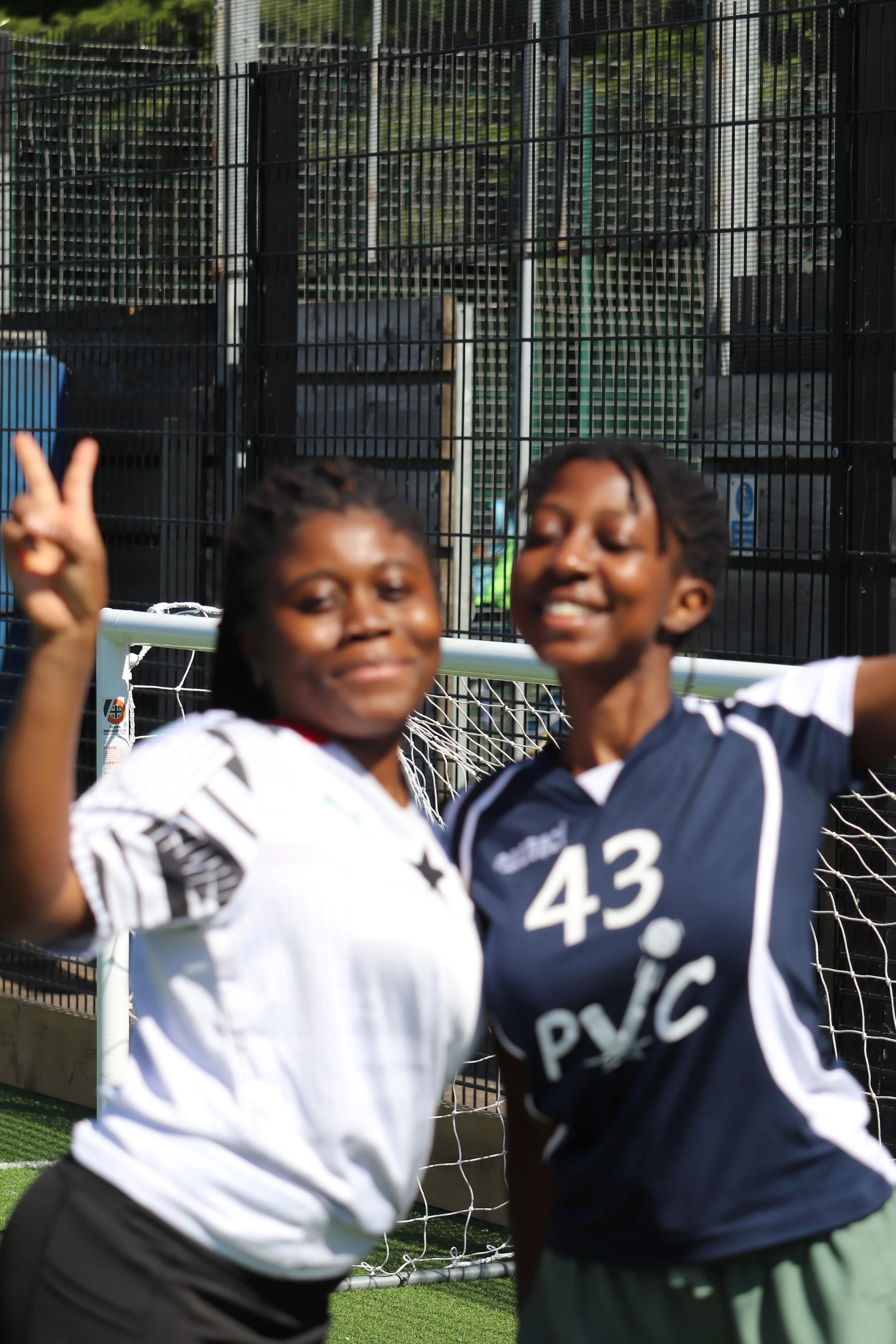 Two young women are smiling and posing happily on a sports field, with a soccer goal and a black metal fence in the background.