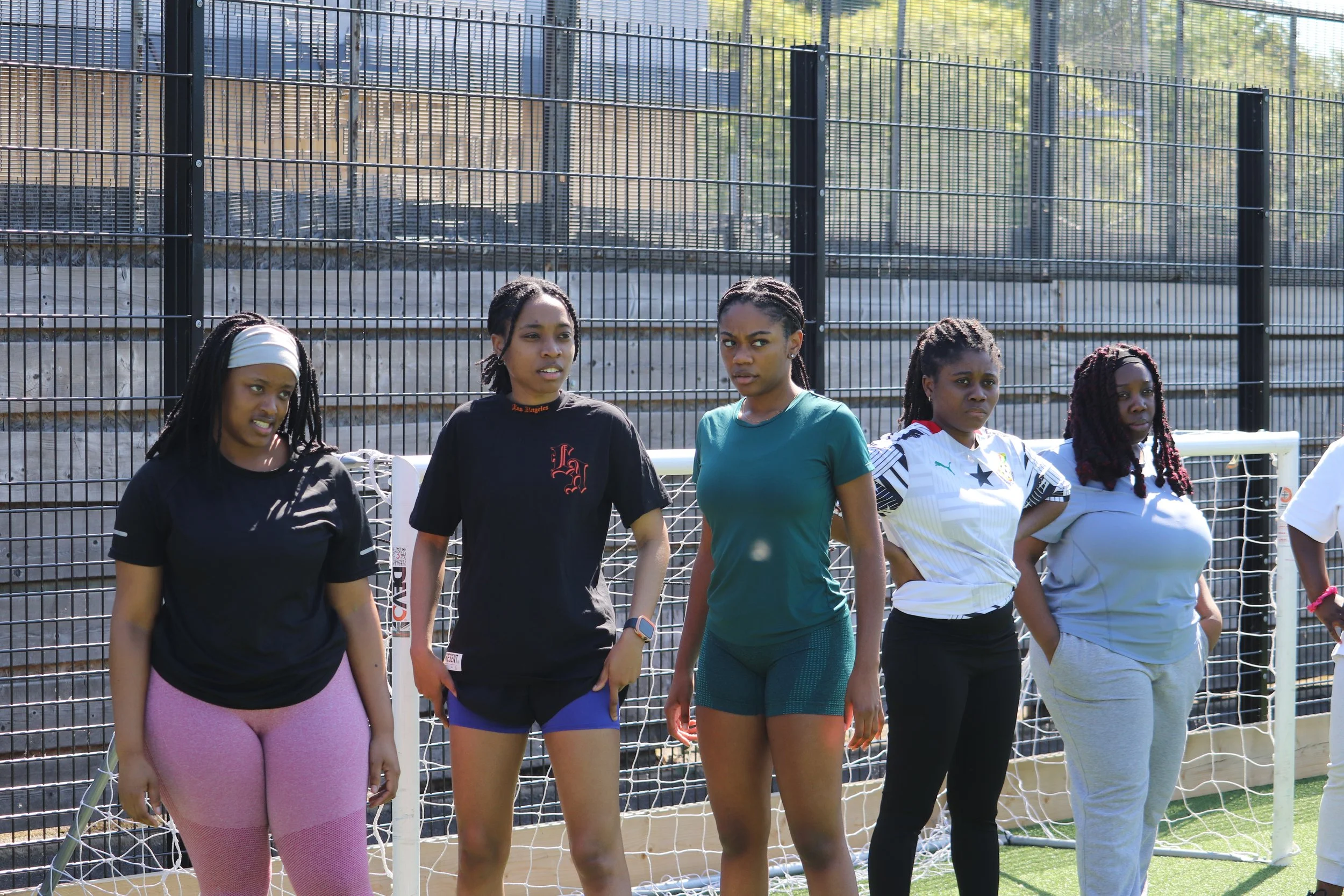 Five women standing on a sports field in front of a metal fence, some with hands on hips, appearing focused and attentive.