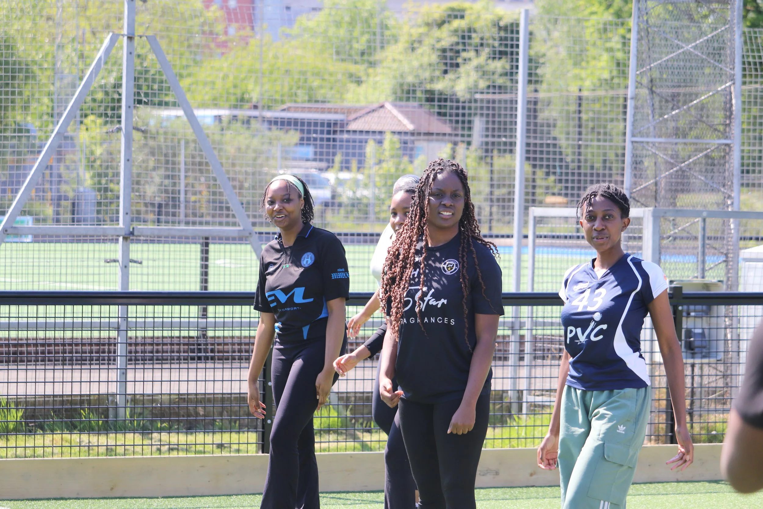 Four women standing on a sports field with a metal fence and green trees in the background, dressed in athletic clothing, some smiling and others with neutral expressions.