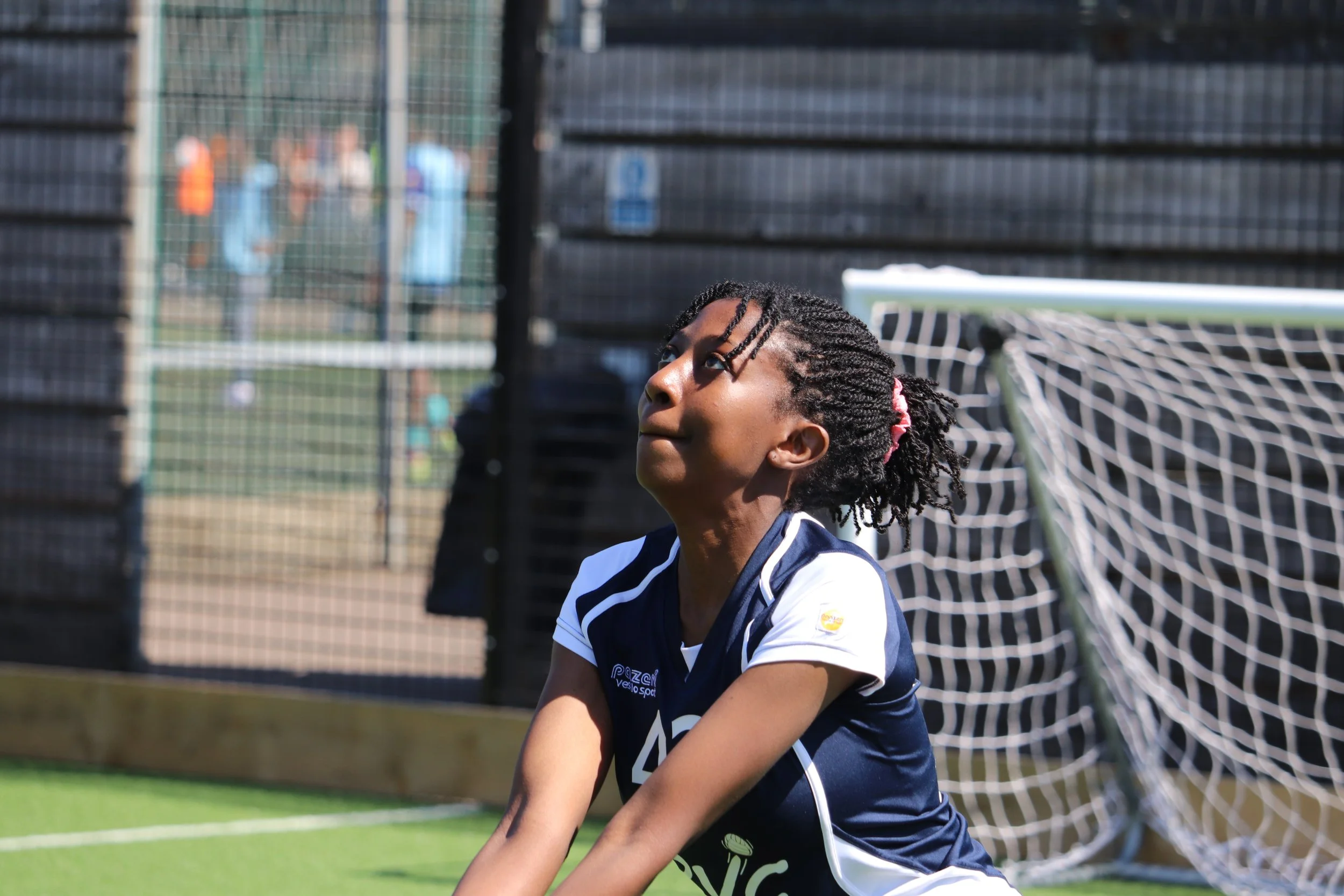 A young girl with braided hair and a pink hair tie, wearing a navy blue and white sports jersey, on a soccer field near a goalpost.
