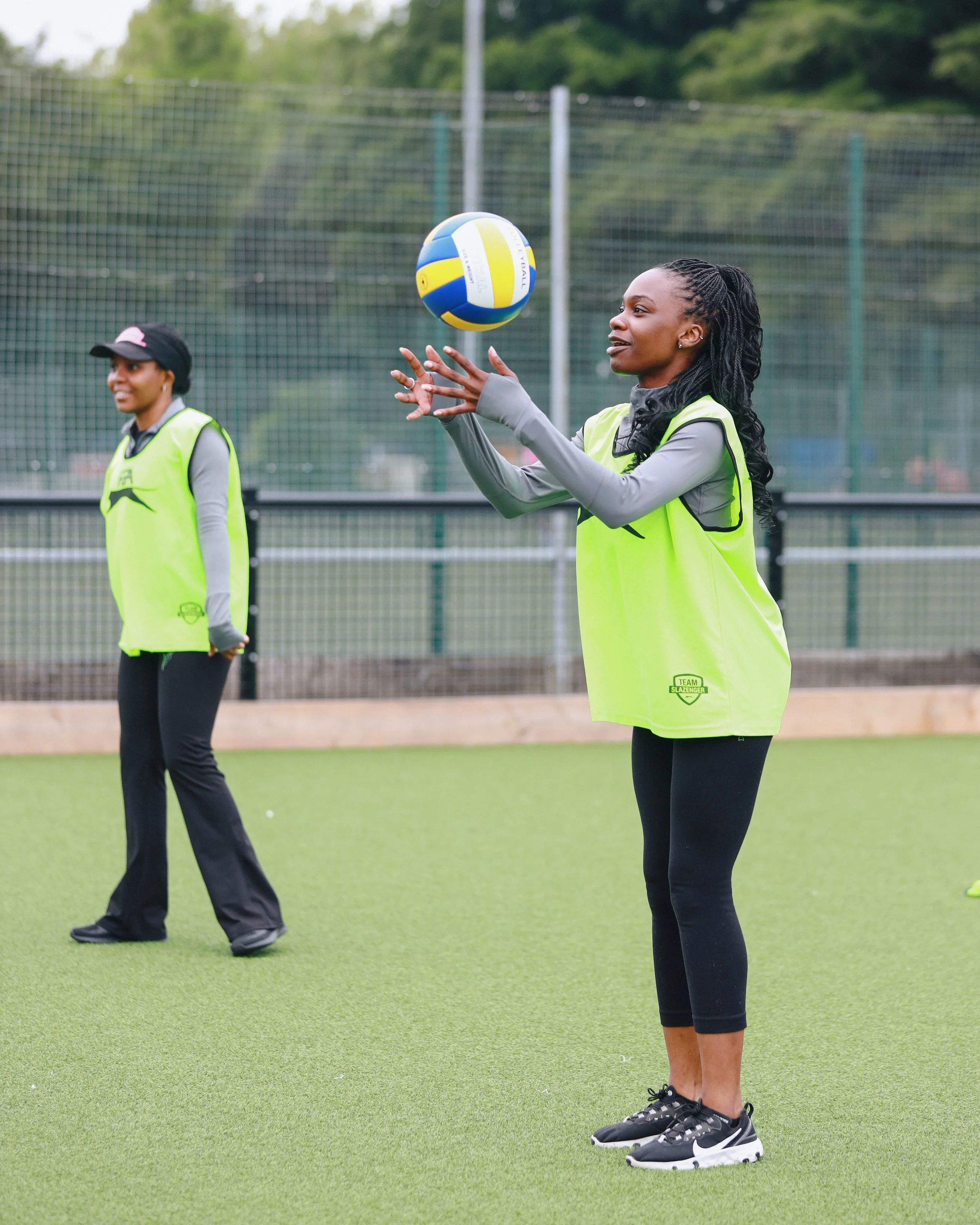 Two women playing volleyball on an outdoor court; one woman in the foreground wearing a neon green vest and black leggings, preparing to catch or serve the ball, and a woman in the background wearing a similar vest and a cap, watching.